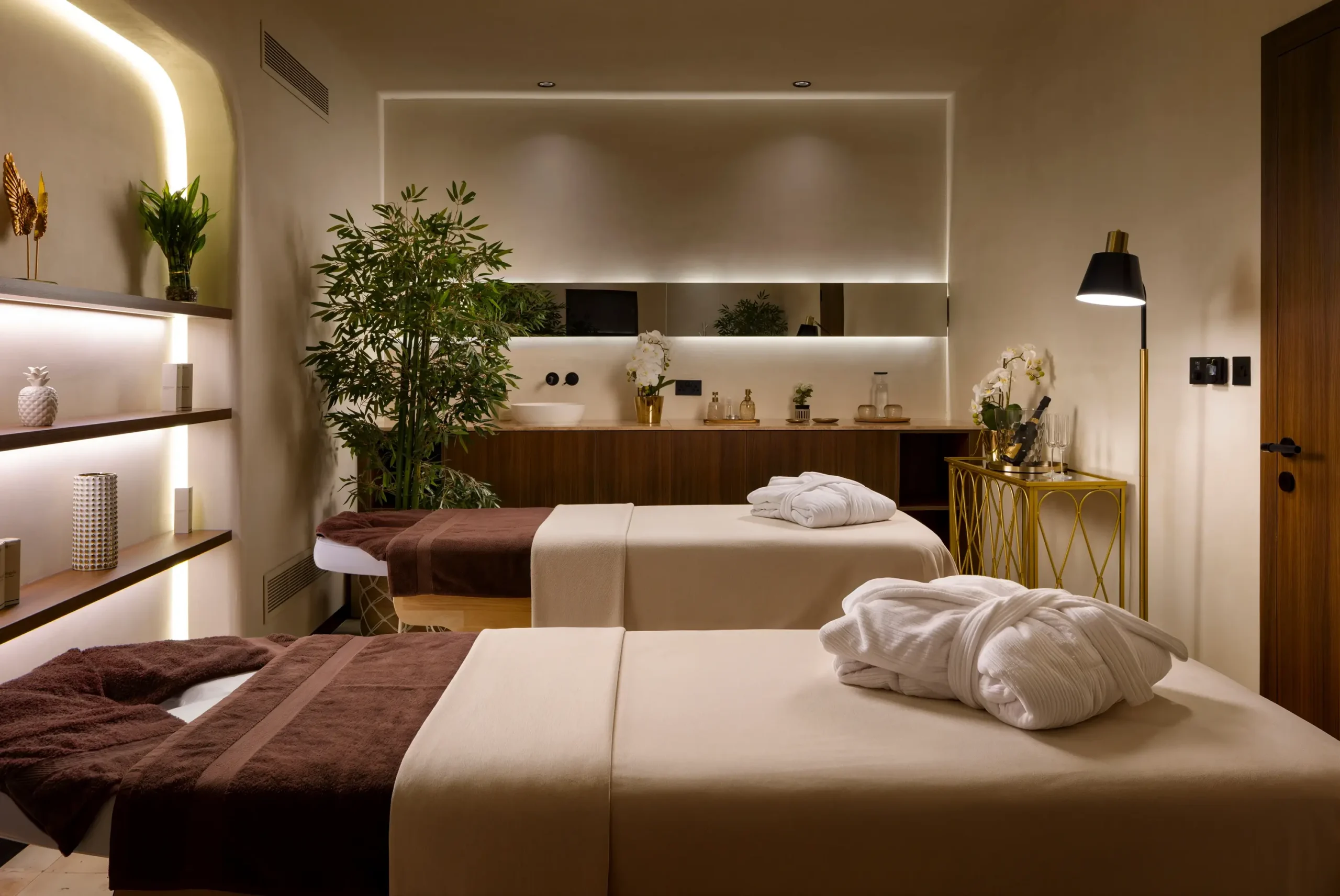Two spa treatment beds with rolled robes in a warm room with lit shelving