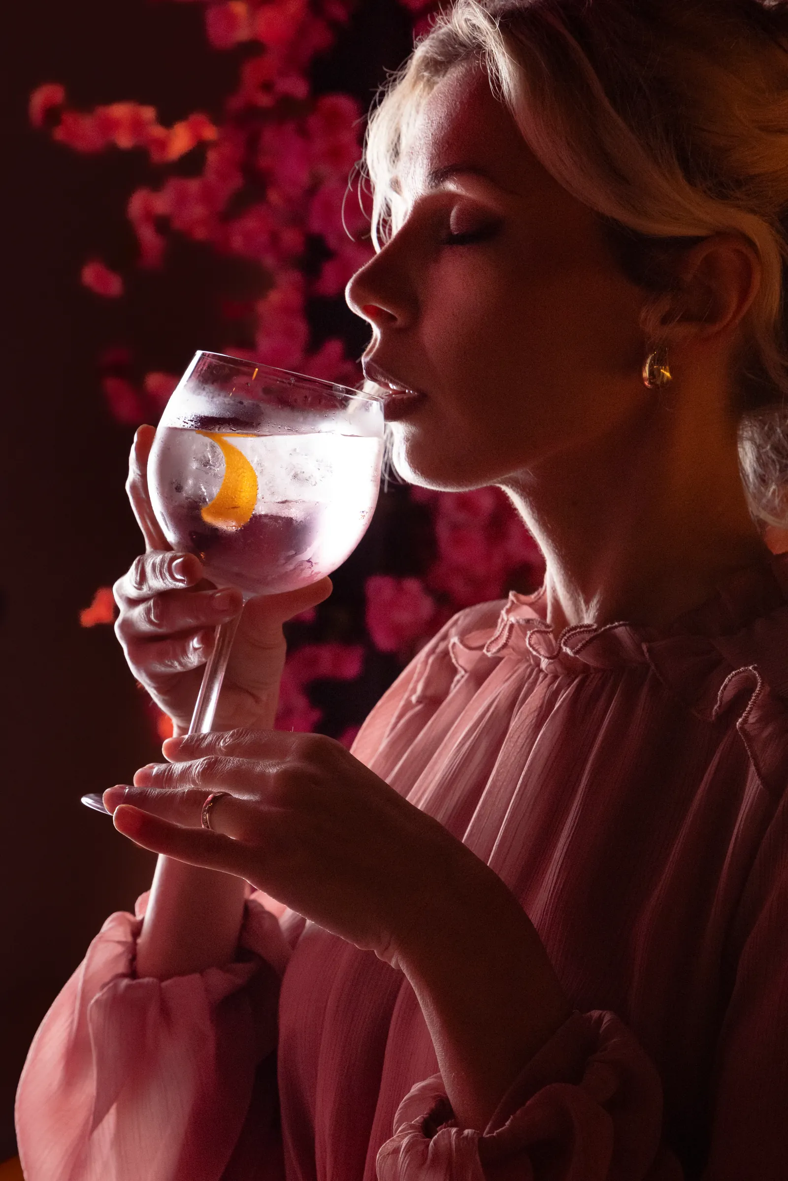 Woman in pink holding a cocktail glass to her nose among blossom