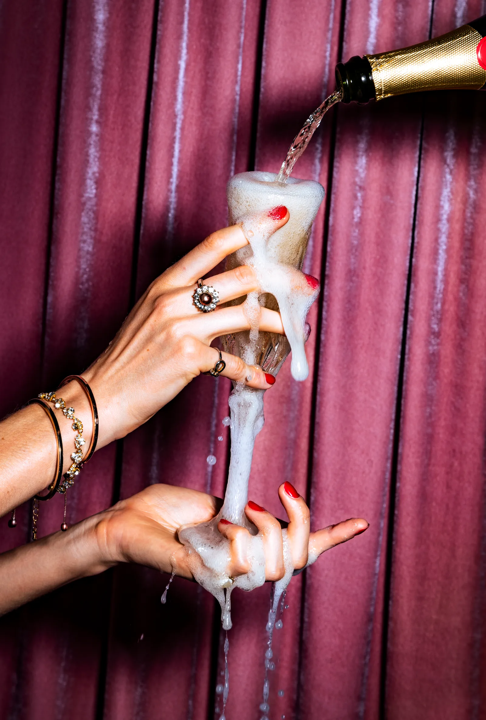 Hands pouring sparkling wine into a glass against a burgundy curtain