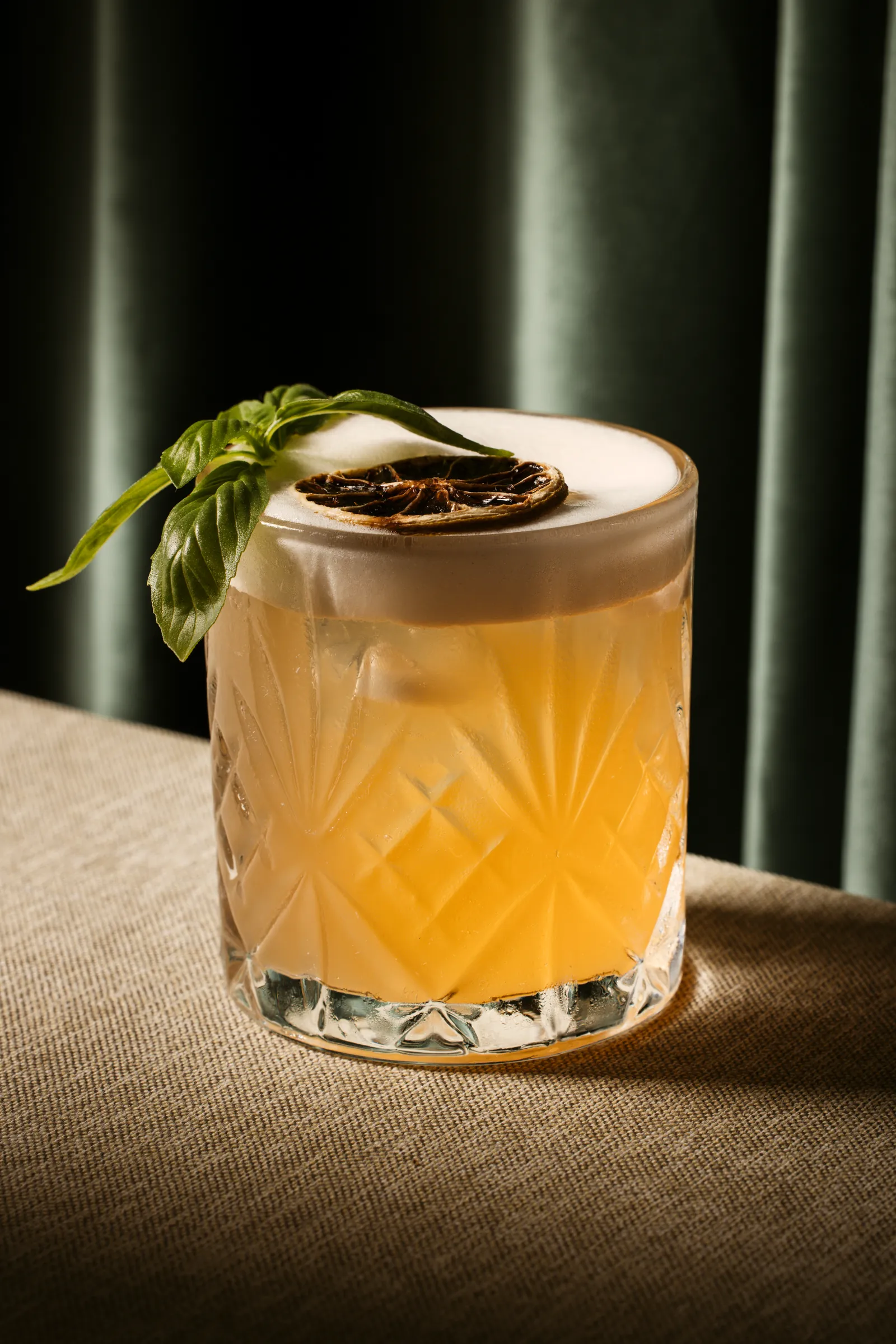 Cocktail with a herb garnish in a cut-crystal glass against a green curtain