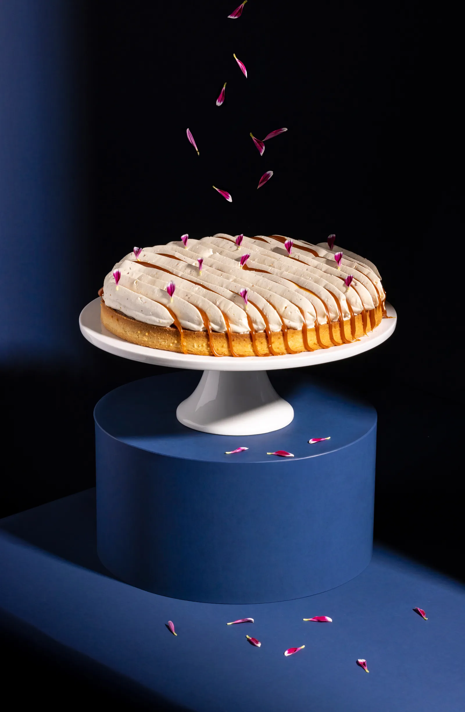 Cake on a stand with rose petals falling against a dark background