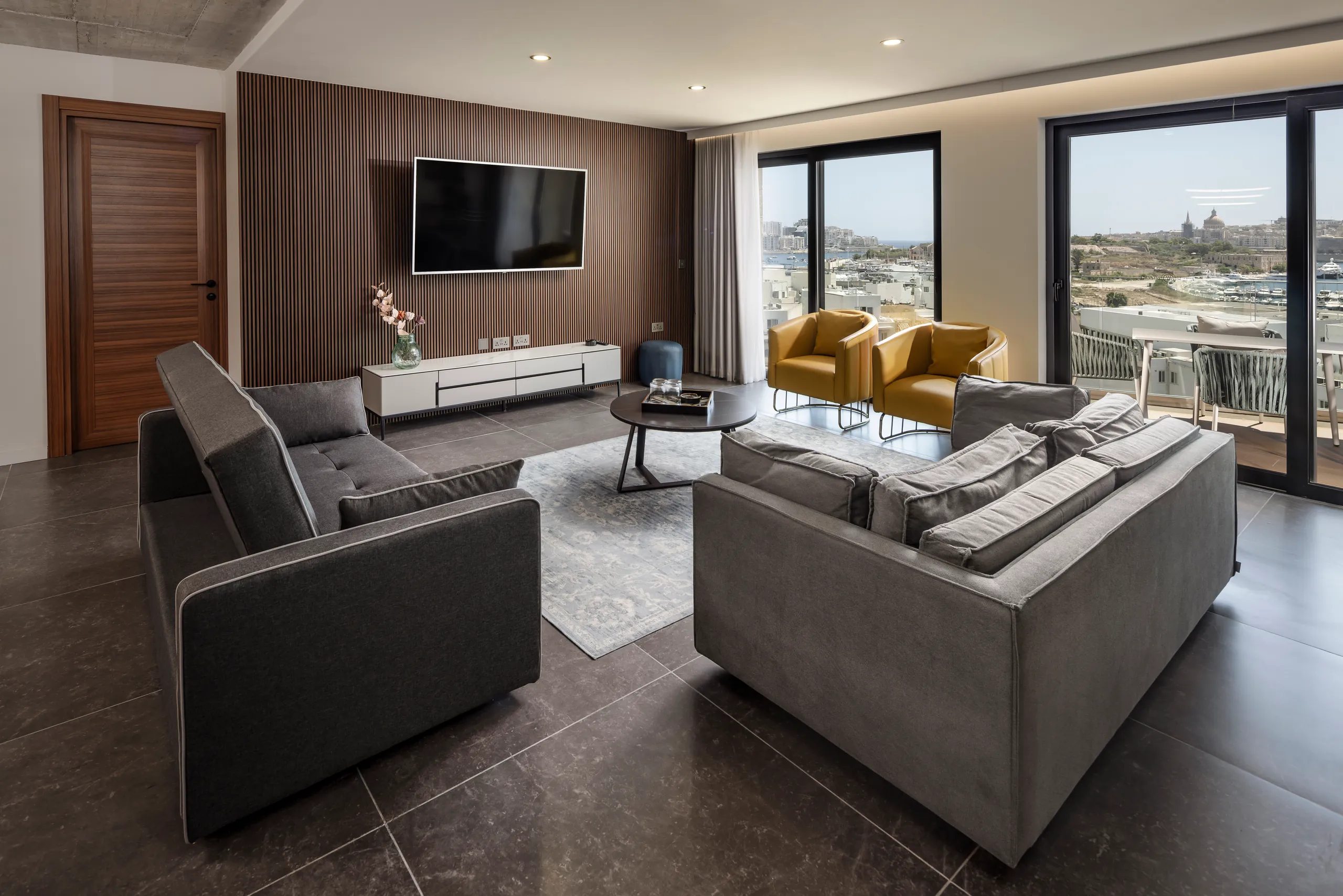 Suite lounge with grey sofas, a wall-mounted TV and a view over rooftops