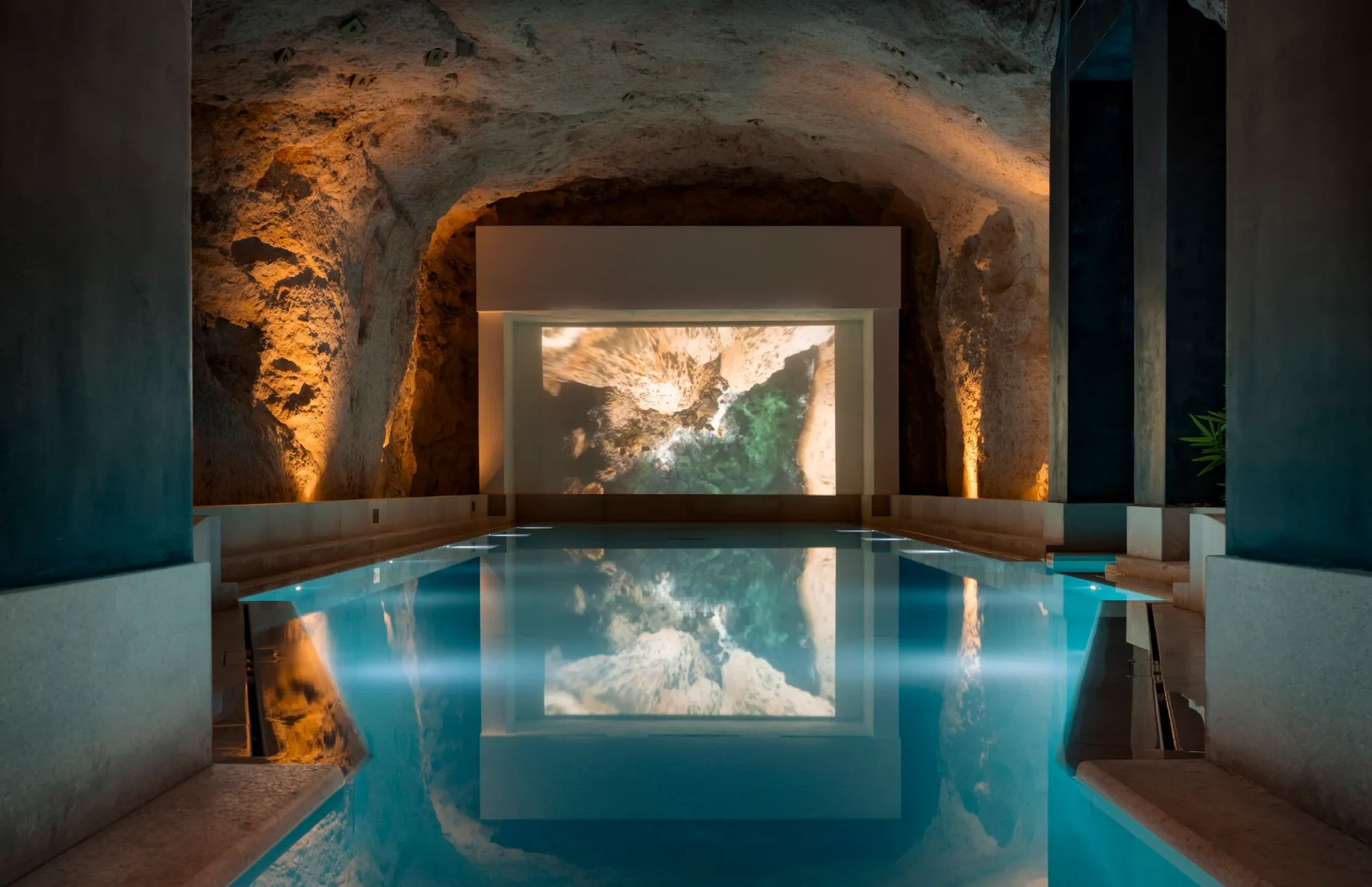 Indoor spa pool in a rock-walled cave setting with a lit screen at the far end