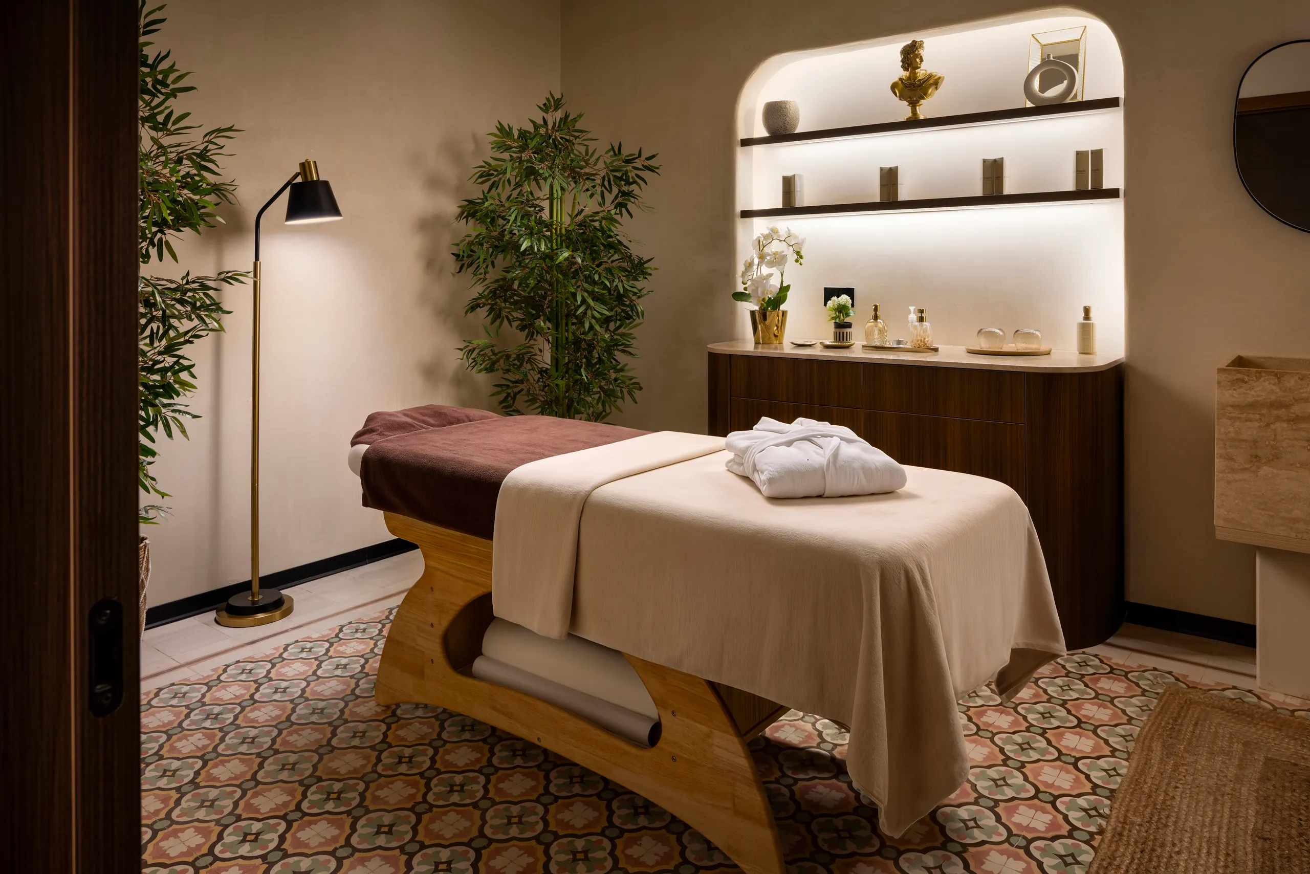 Spa treatment room with a massage bed, folded robe, patterned floor tiles and lit shelves