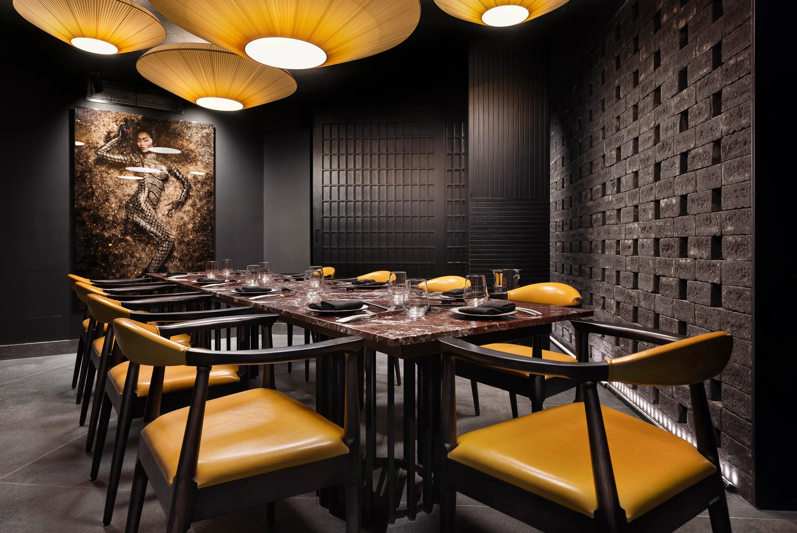 Dining table with mustard chairs under woven pendants, in front of a carved stone relief