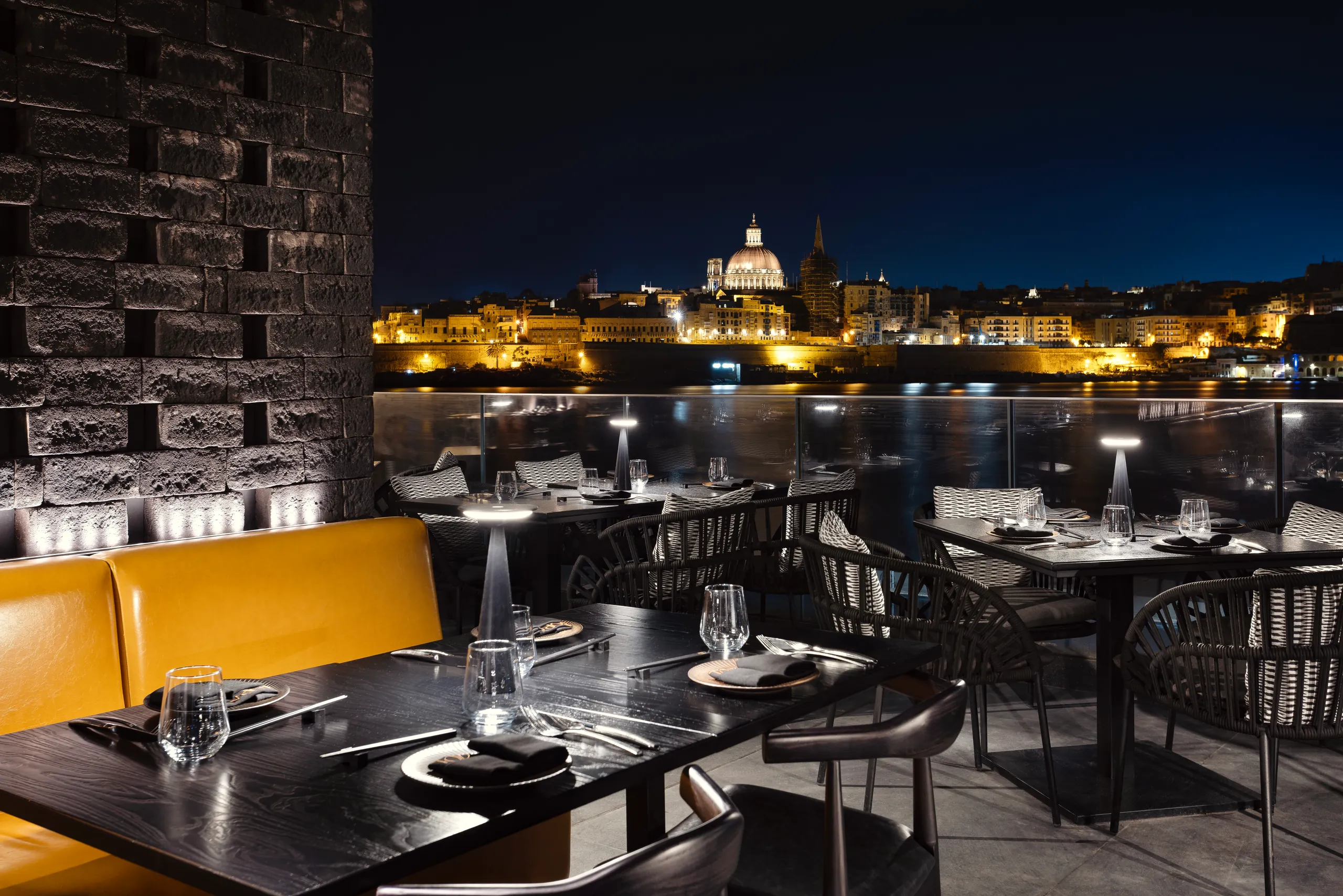 Terrace table at night with a mustard banquette and the floodlit Valletta skyline beyond