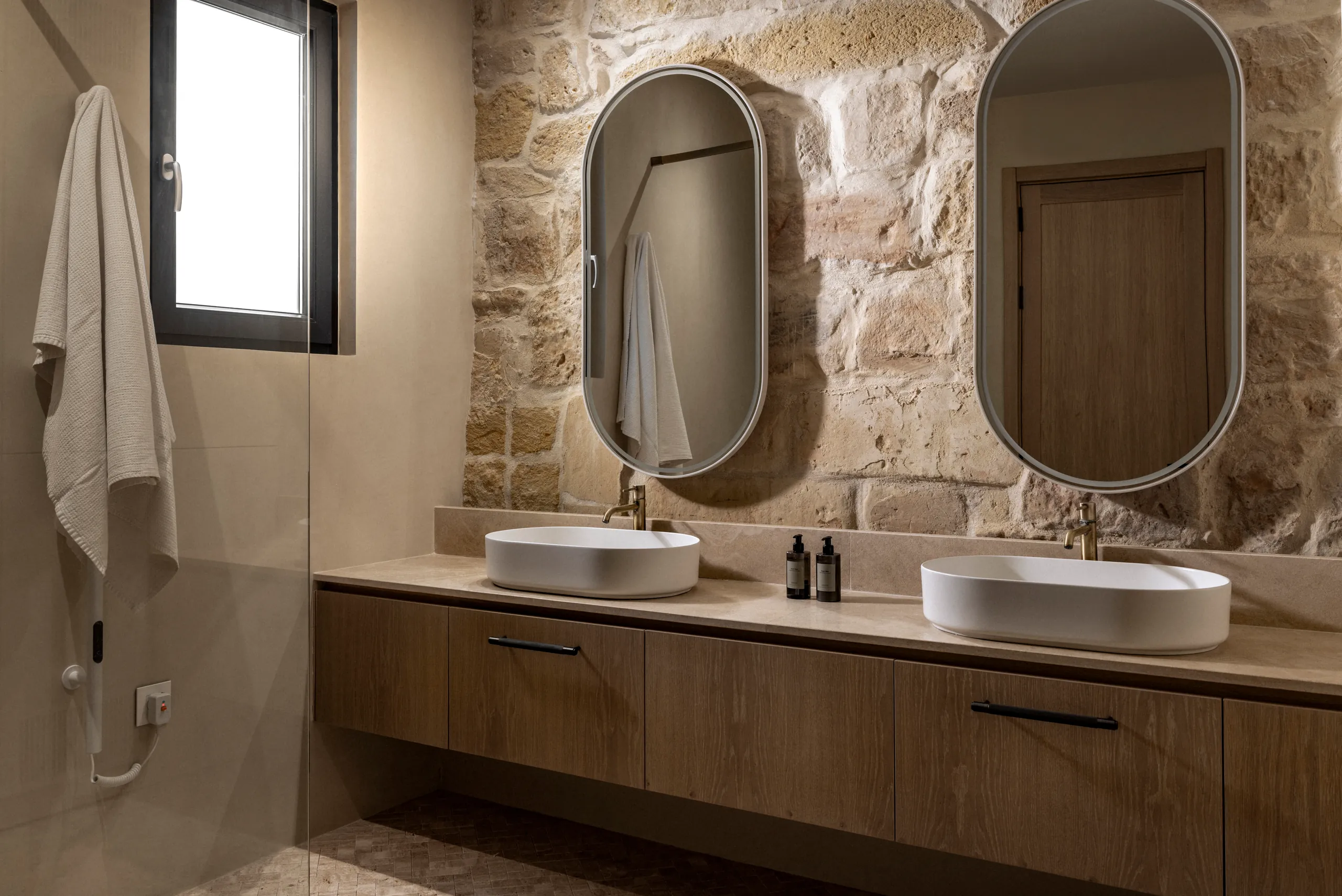 Bathroom with two oval mirrors, twin basins, timber vanity and a limestone wall
