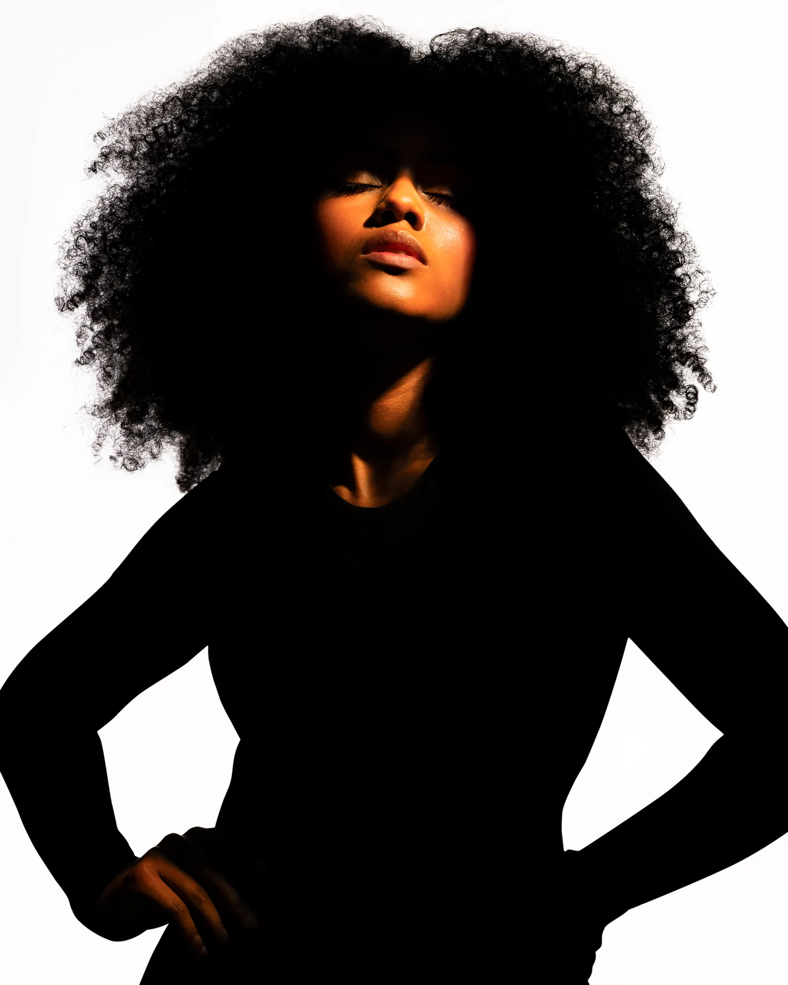 Woman with voluminous natural hair in dramatic backlight, hands on hips, in black