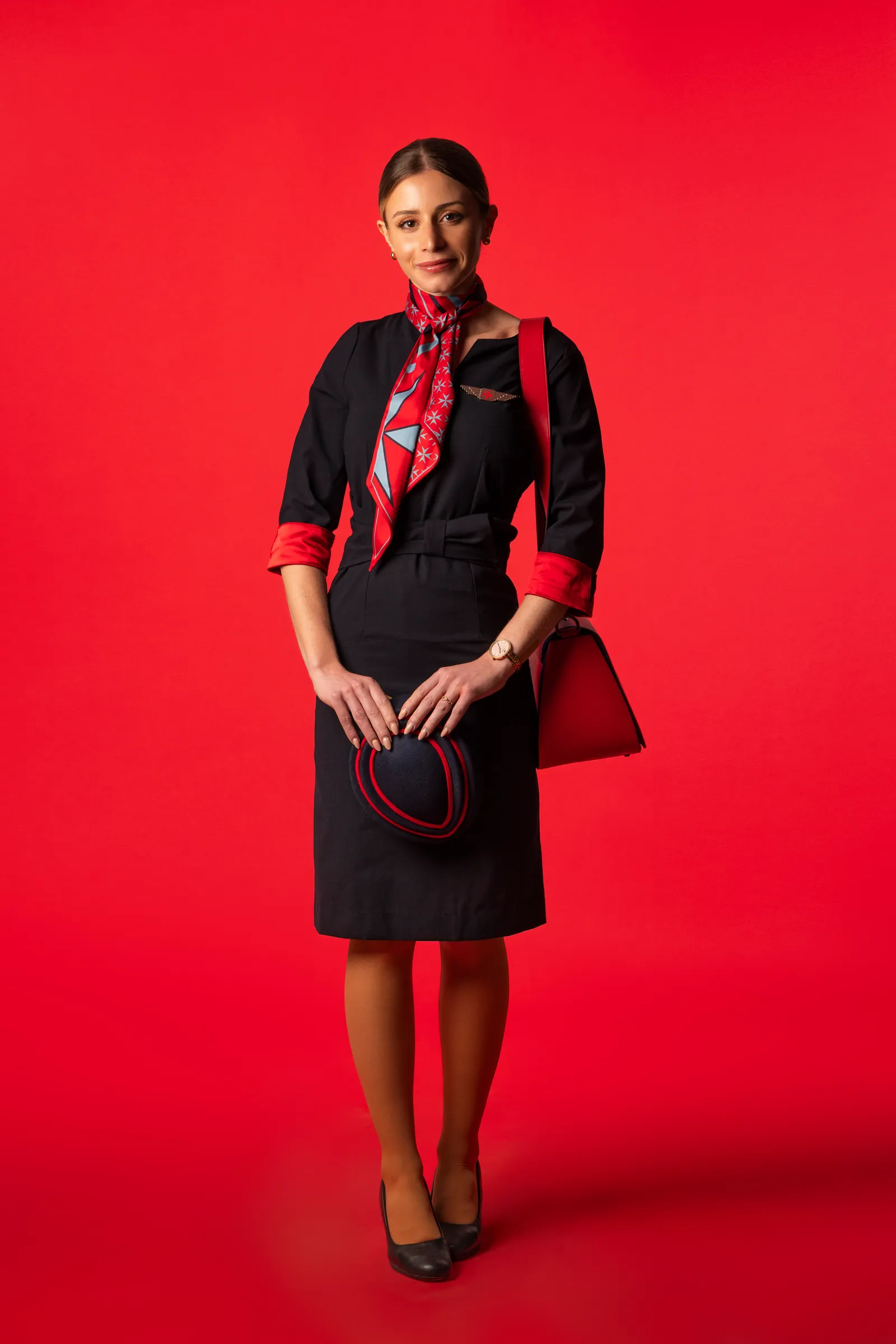 KM Malta Airlines flight attendant in a navy uniform and Charles & Ron scarf against a red background