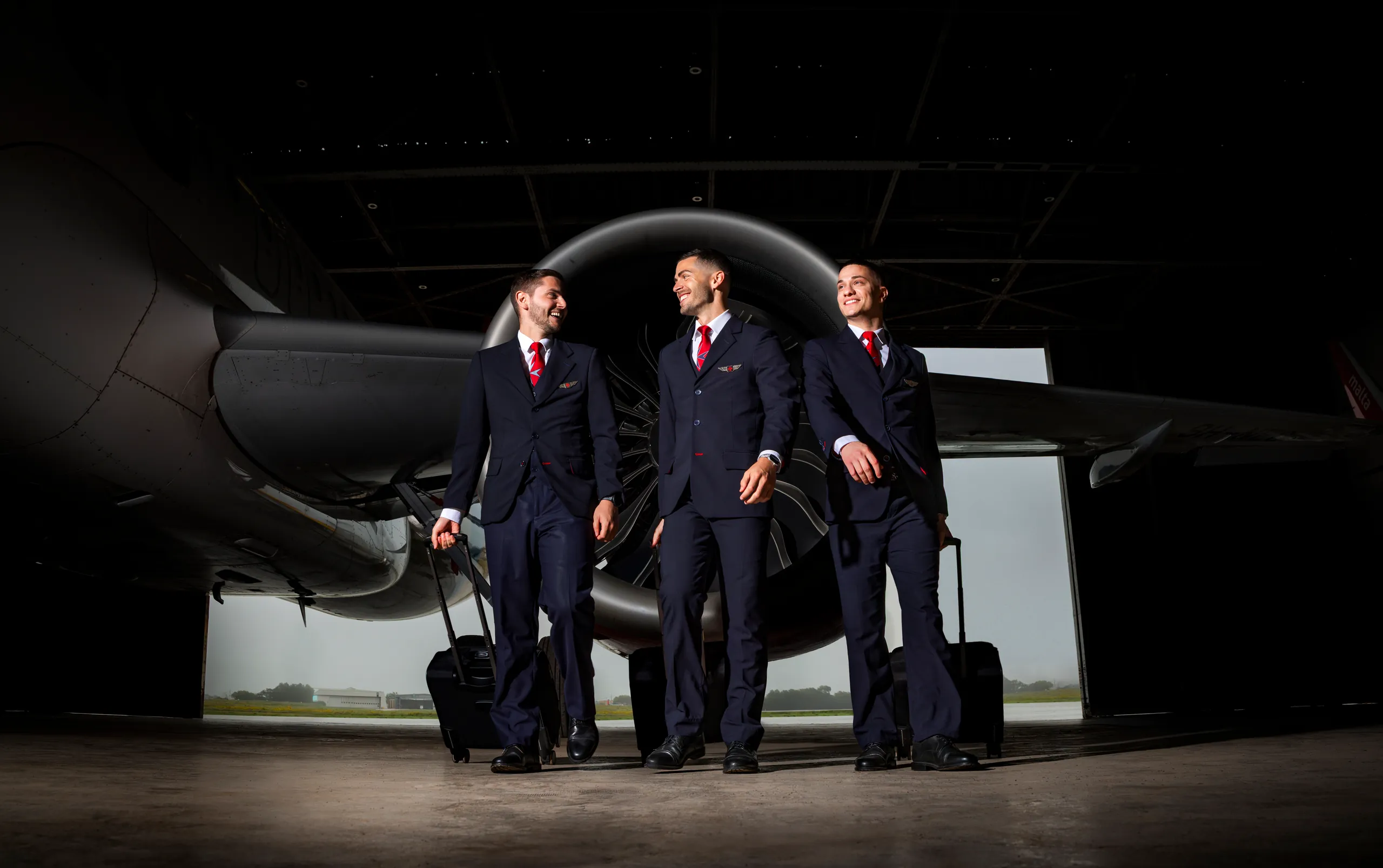 KM Malta Airlines pilots and crew in navy suits with red ties wheeling luggage past a jet engine