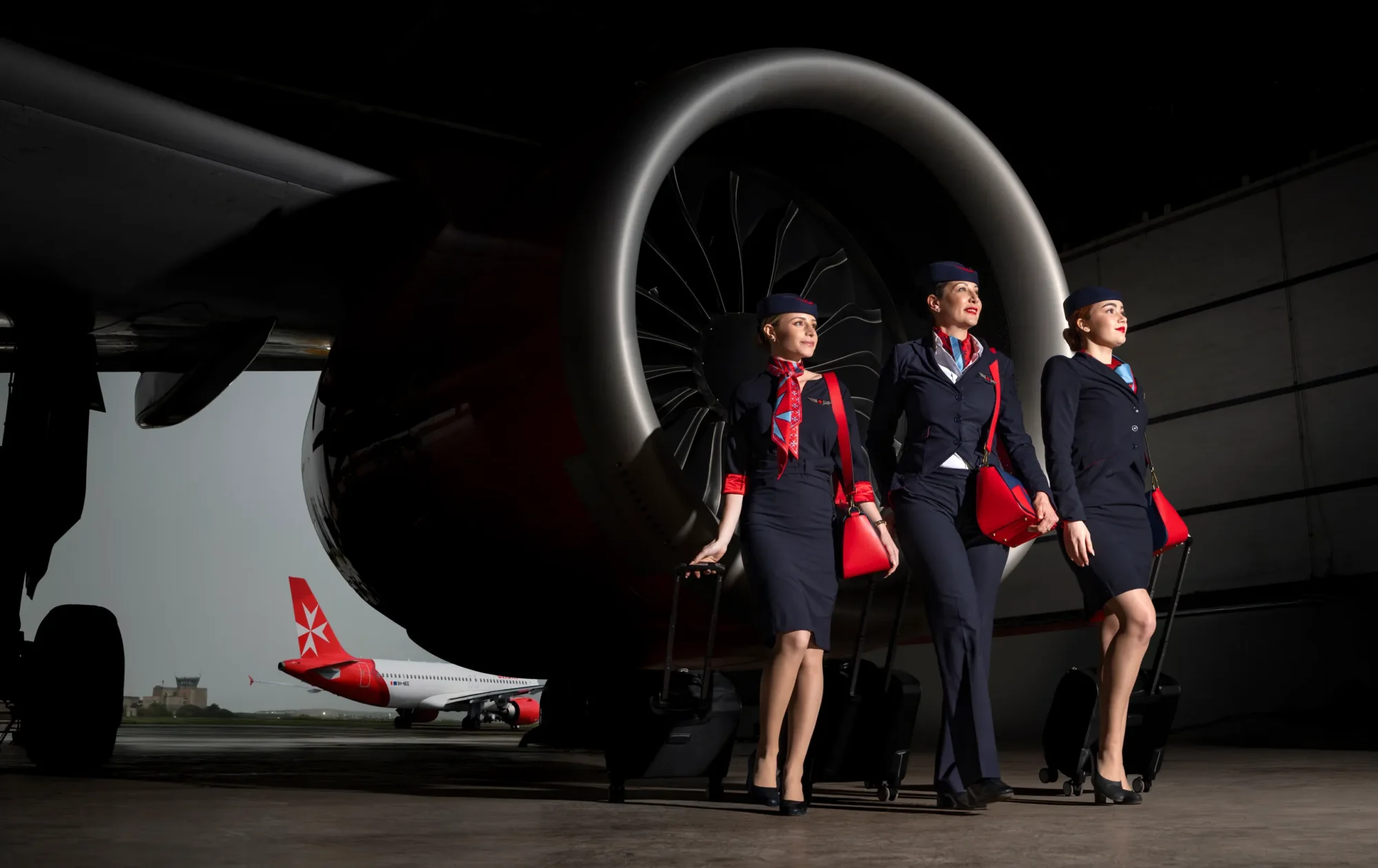 KM Malta Airlines cabin crew in navy uniforms before a jet engine, with a red-tailed aircraft behind