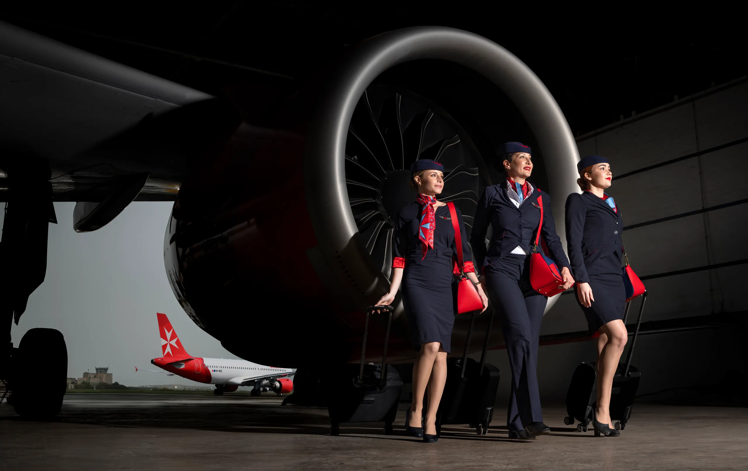 KM Malta Airlines cabin crew in navy uniforms before a jet engine, with a red-tailed aircraft behind