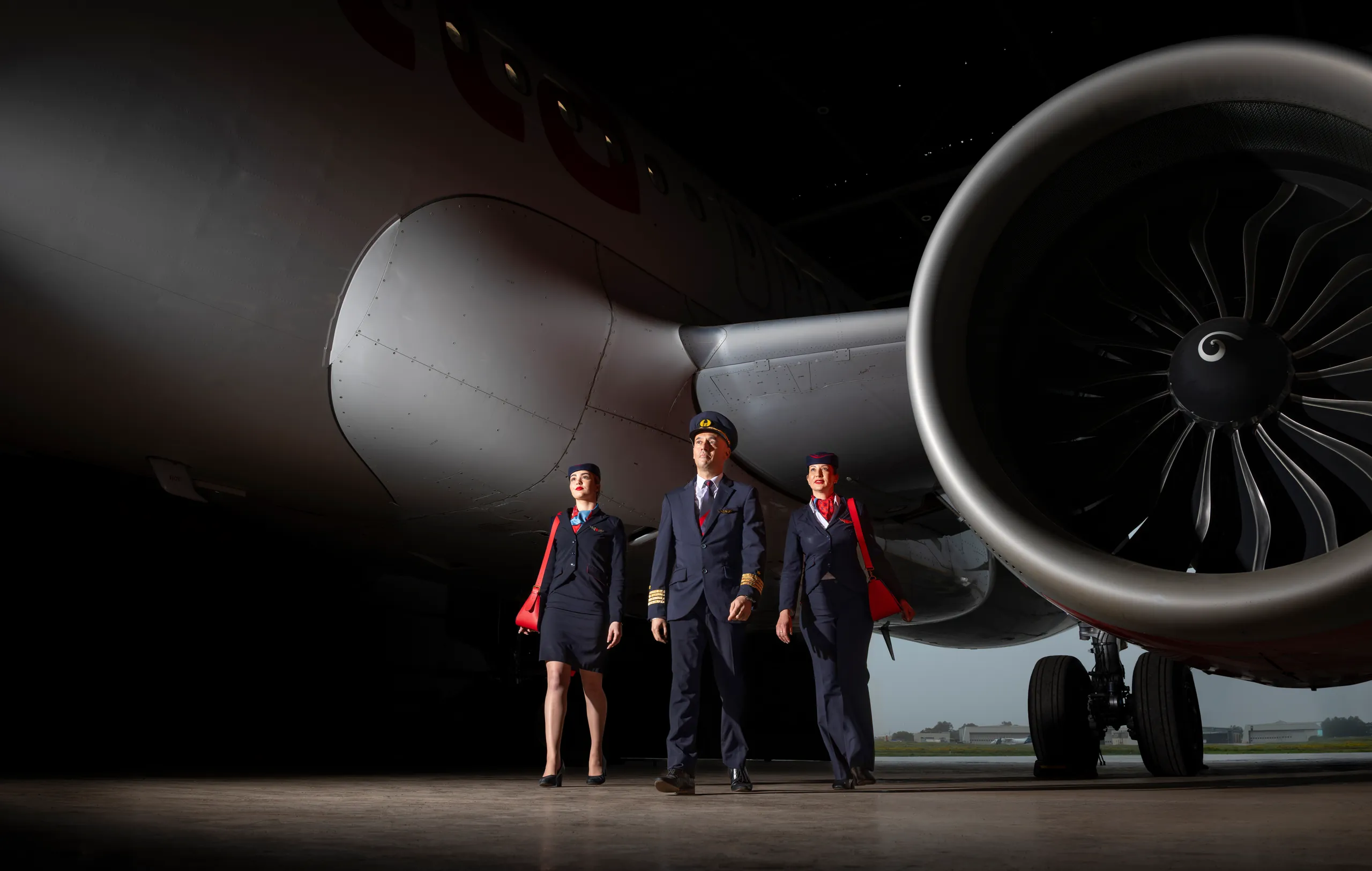 KM Malta Airlines pilot and two cabin crew walking towards camera beneath a jet engine at dusk
