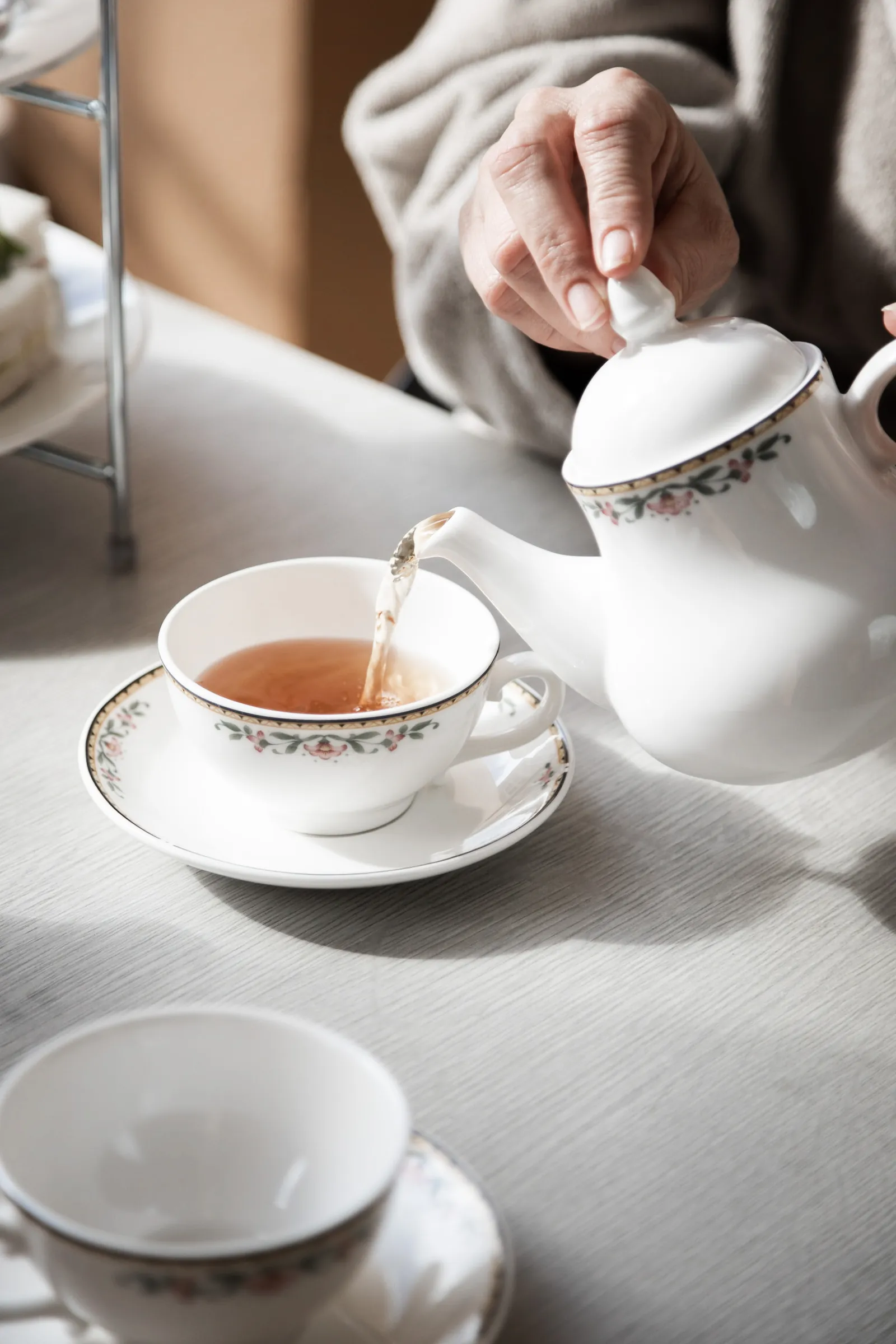 Hand pouring tea at a white table beside a cup and saucer