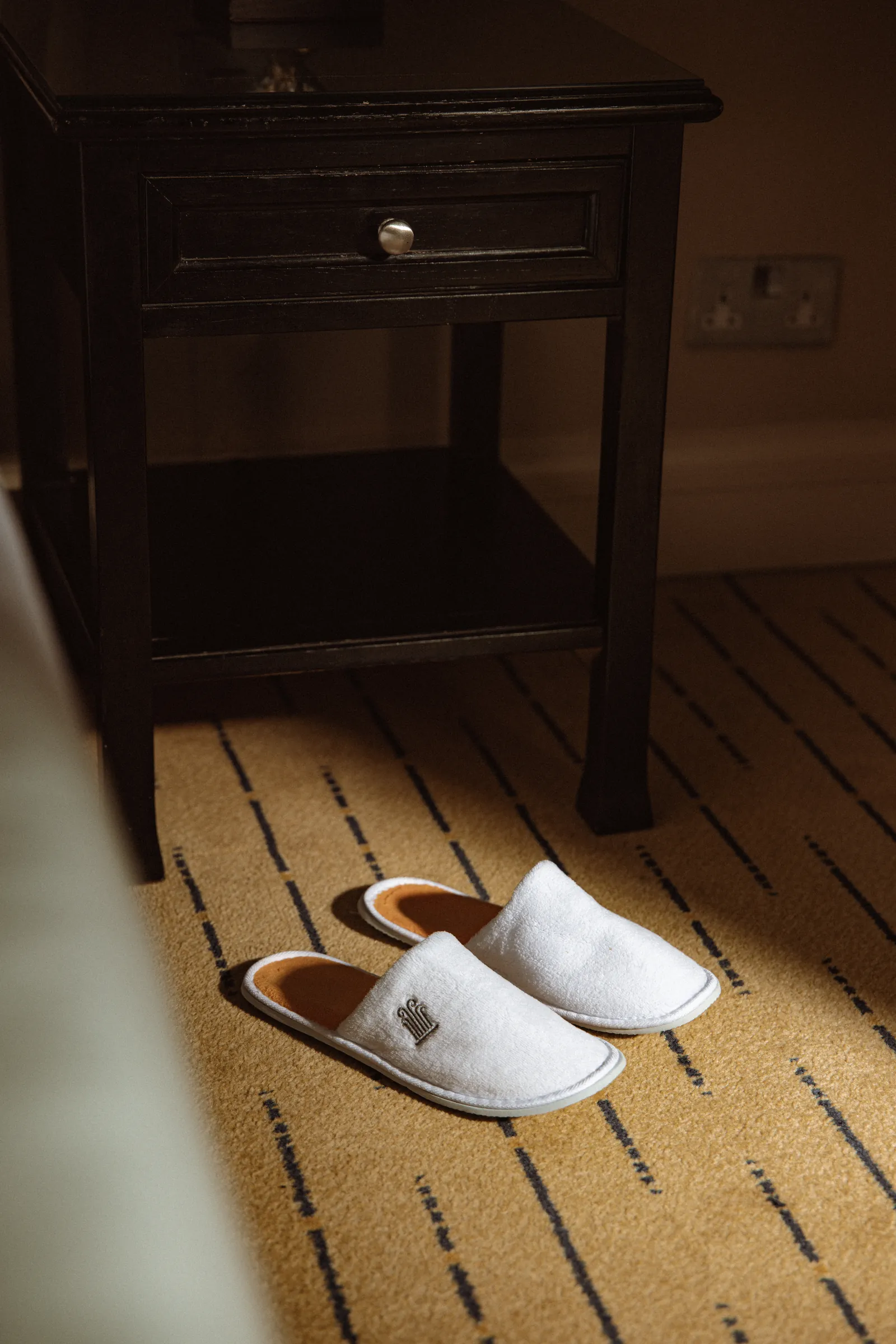 White slippers on the carpet beside a bedside table in a Corinthia guest room