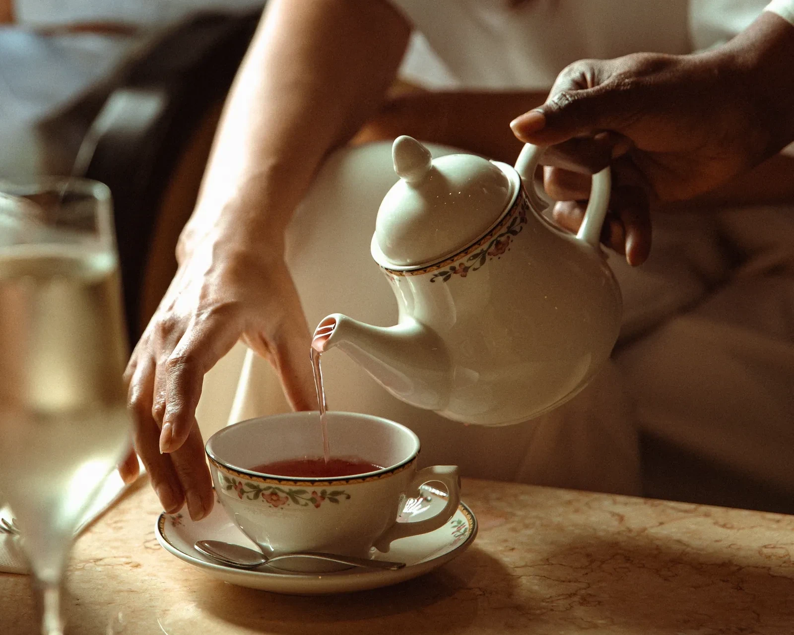 Tea poured from a porcelain teapot into a cup beside a glass at afternoon tea