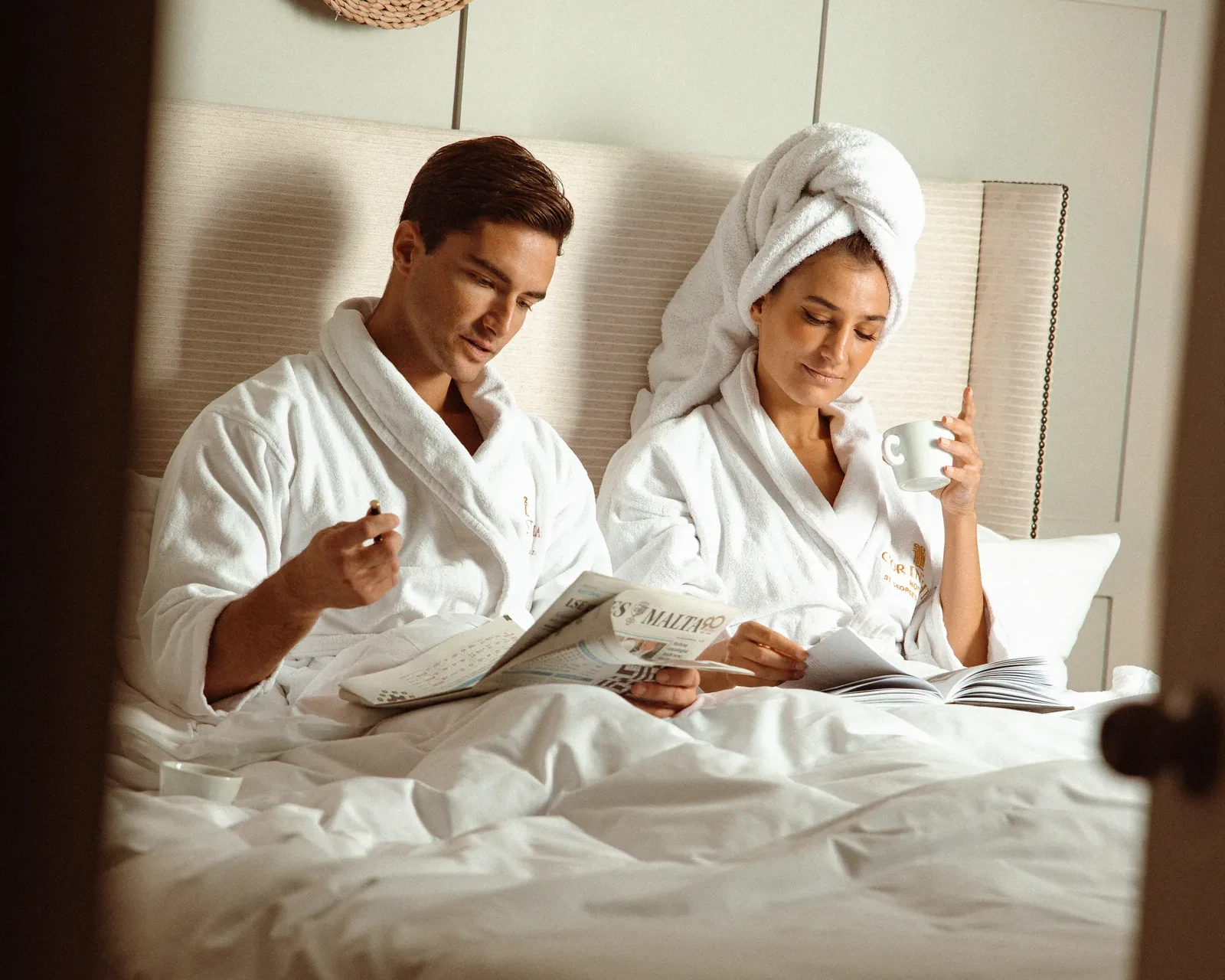 Couple in white robes reading a newspaper and a magazine in bed with tea