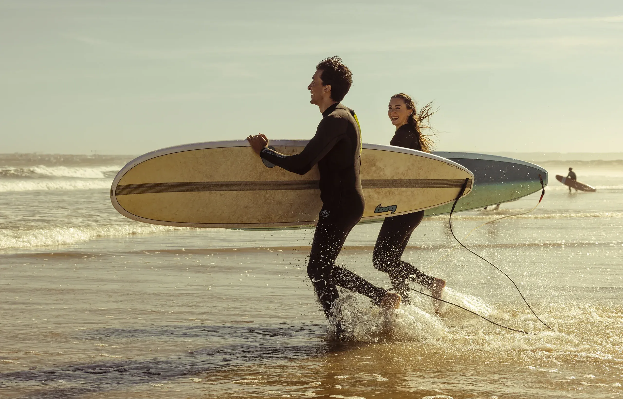 Couple running out of the sea with a surfboard in Portugal, travel photography for Verdi Hotels