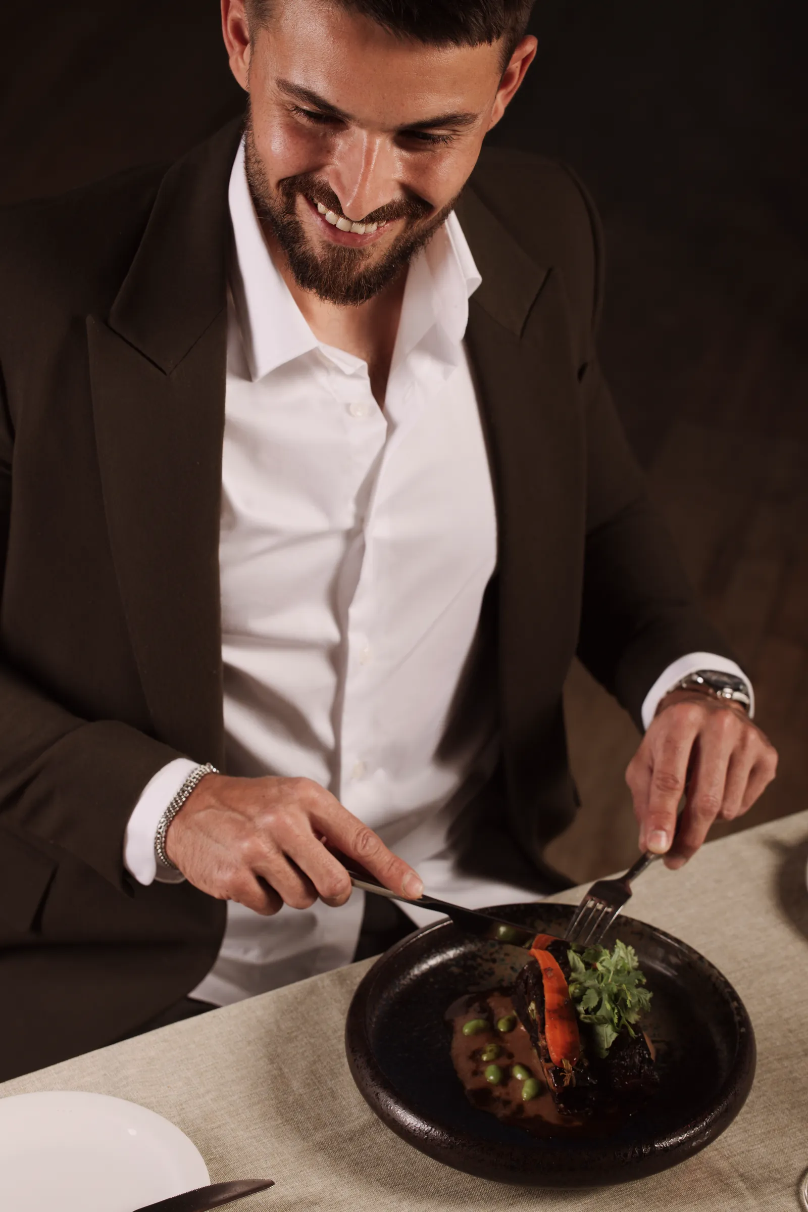 Smiling man in a dark jacket cutting into a plated dish
