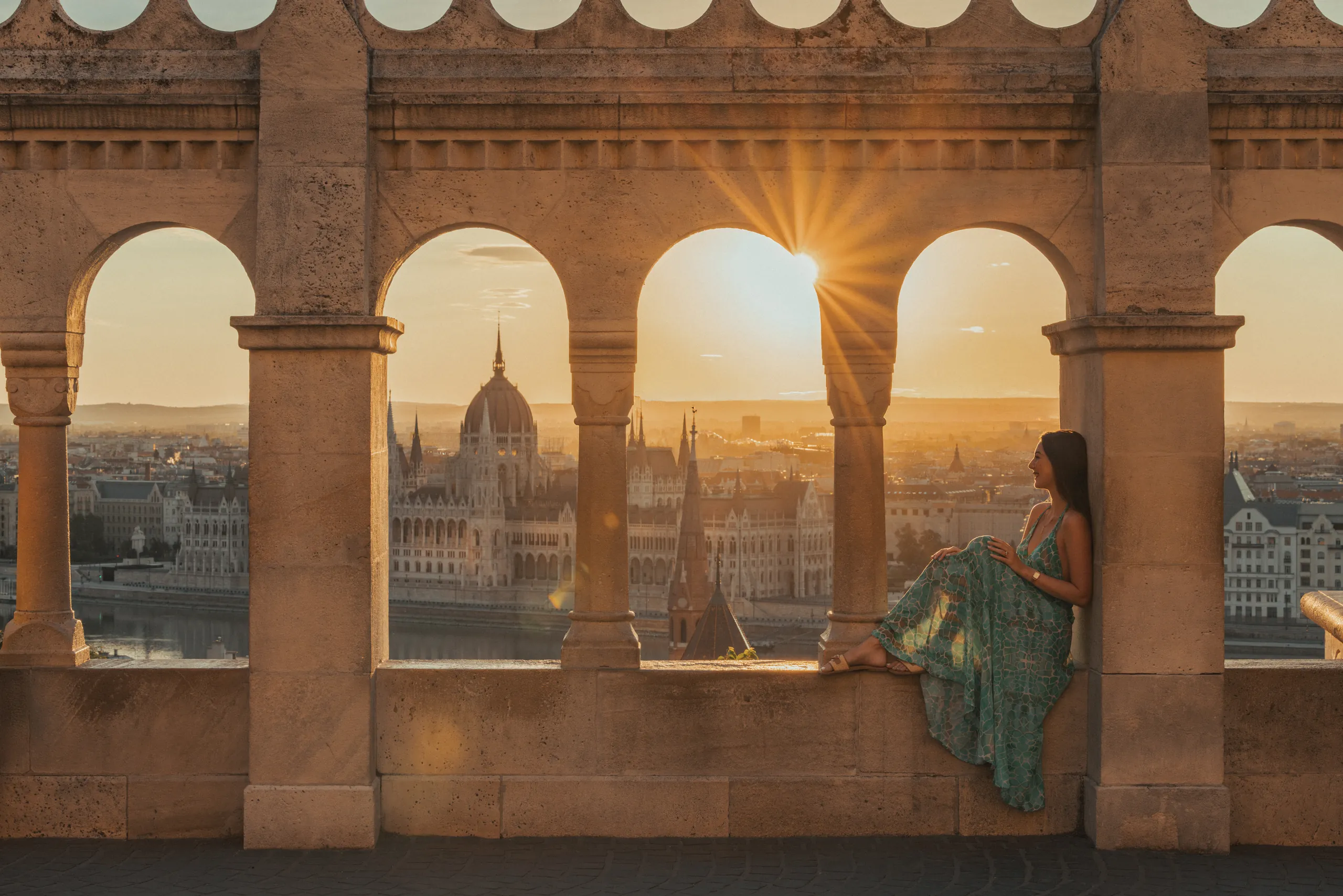 Woman sitting in the arches of Fisherman’s Bastion at sunset, with the Hungarian Parliament Building behind her, Budapest