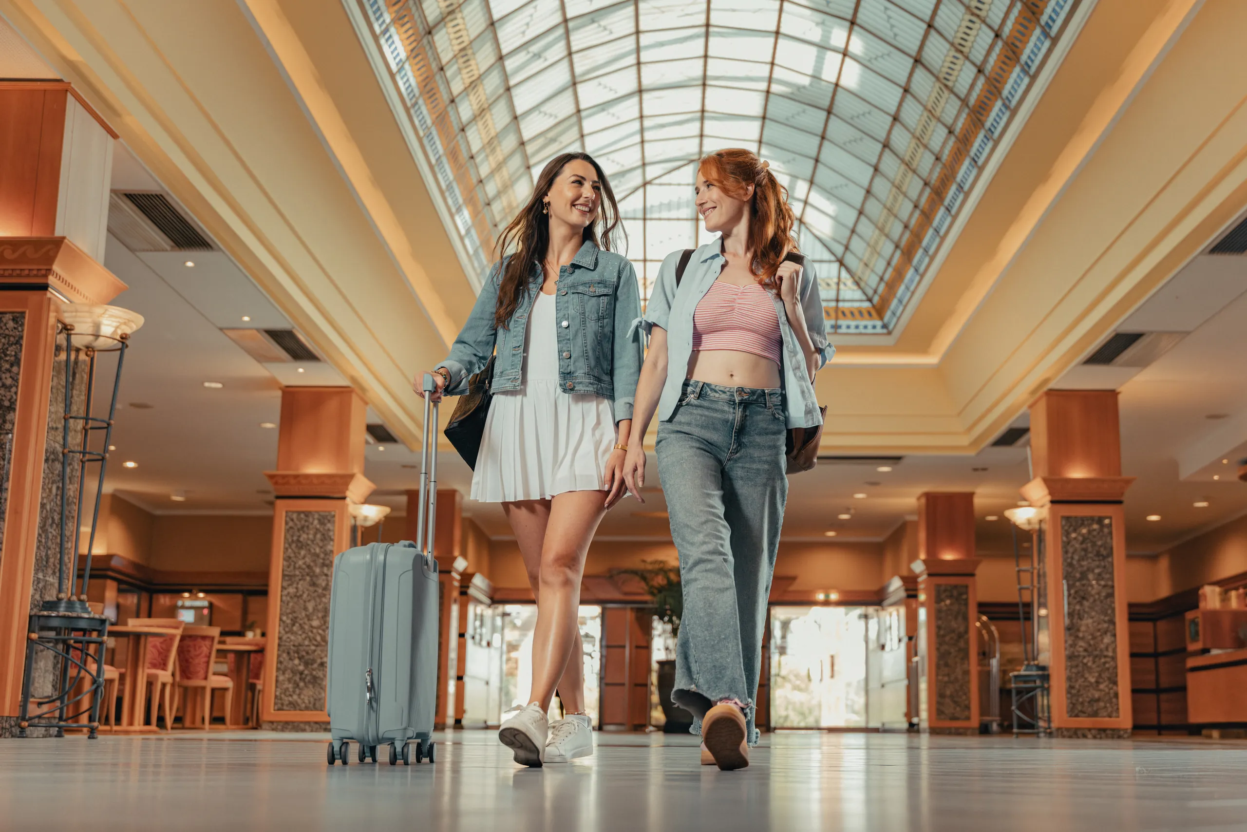 Two friends with luggage walking through the glass-roofed lobby of a hotel in Budapest for Verdi Hotels