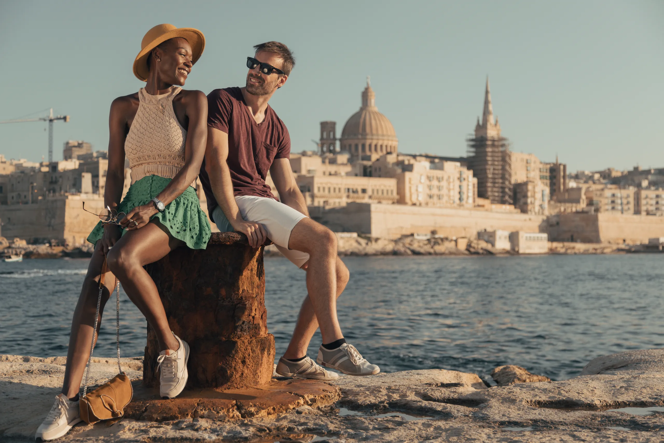Couple sitting on a tree stump on the Malta waterfront with the Valletta skyline behind them, Verdi Hotels, Corinthia Group