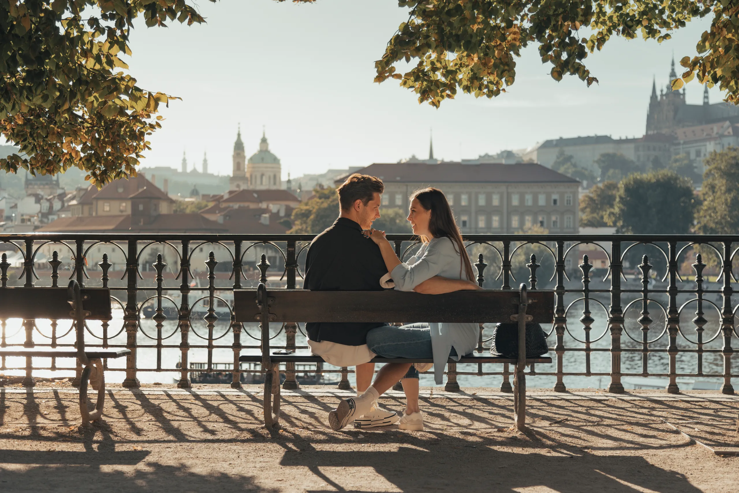 Couple sitting together on a riverside bench in Prague in warm light, with a domed church and Prague Castle in the distance