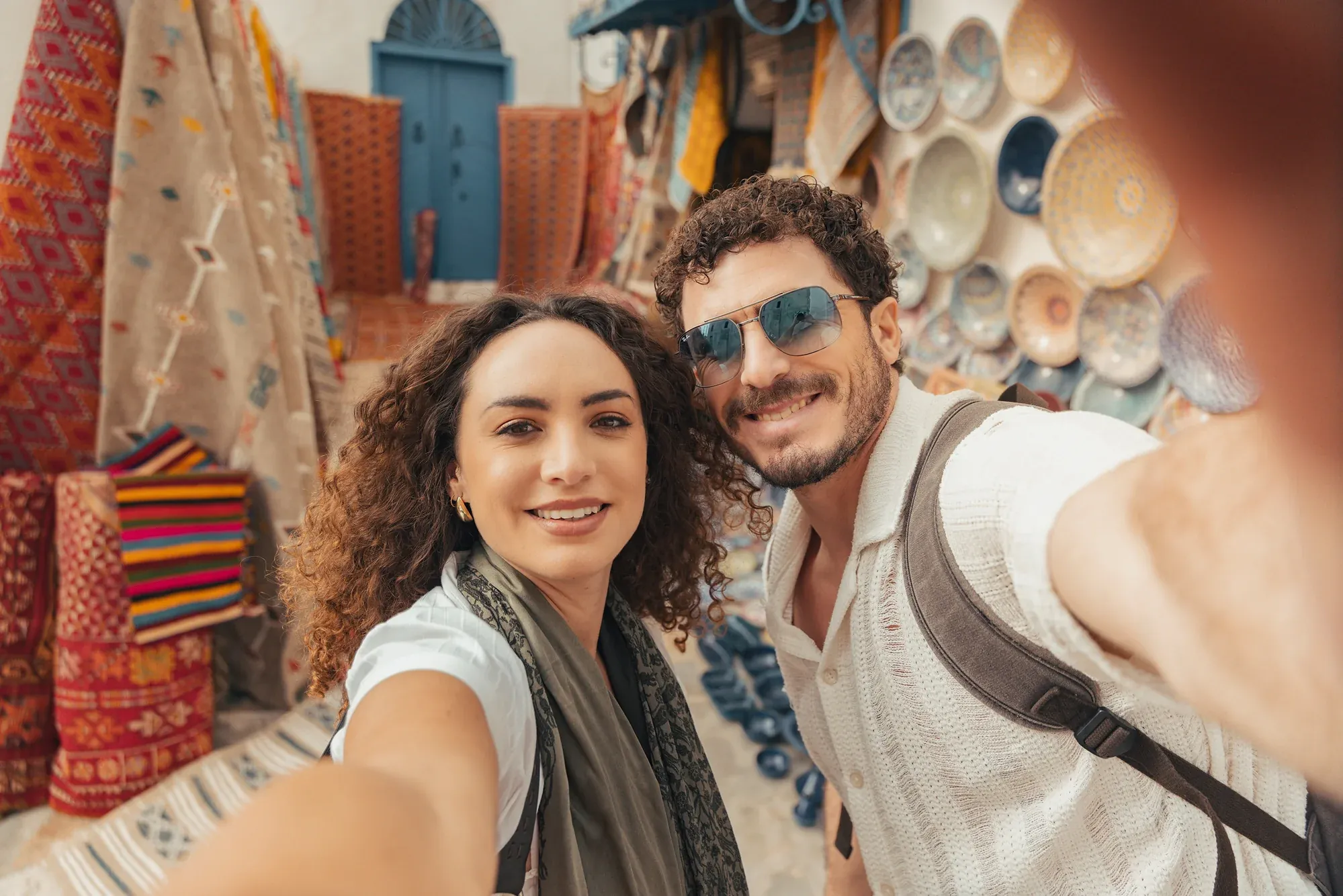 Couple taking a selfie in a souk in Tunisia, with rugs and ceramic plates behind them