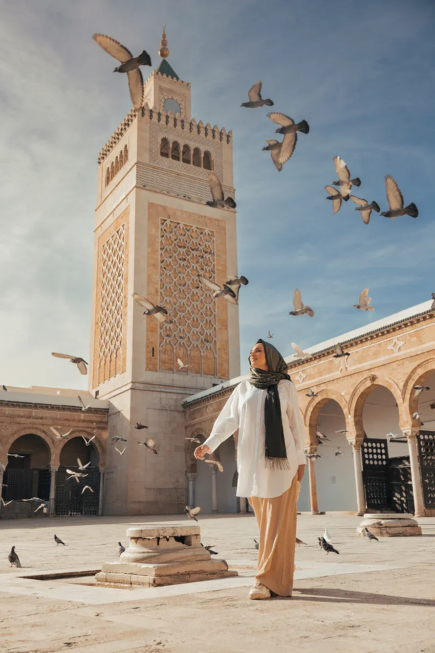 Woman in a white shirt and headscarf standing in a mosque courtyard in Tunis as pigeons fly around the minaret