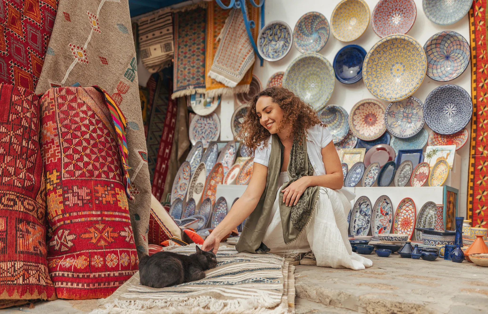 Woman sitting among colourful rugs and ceramic plates in a souk in Tunisia, with a cat beside her