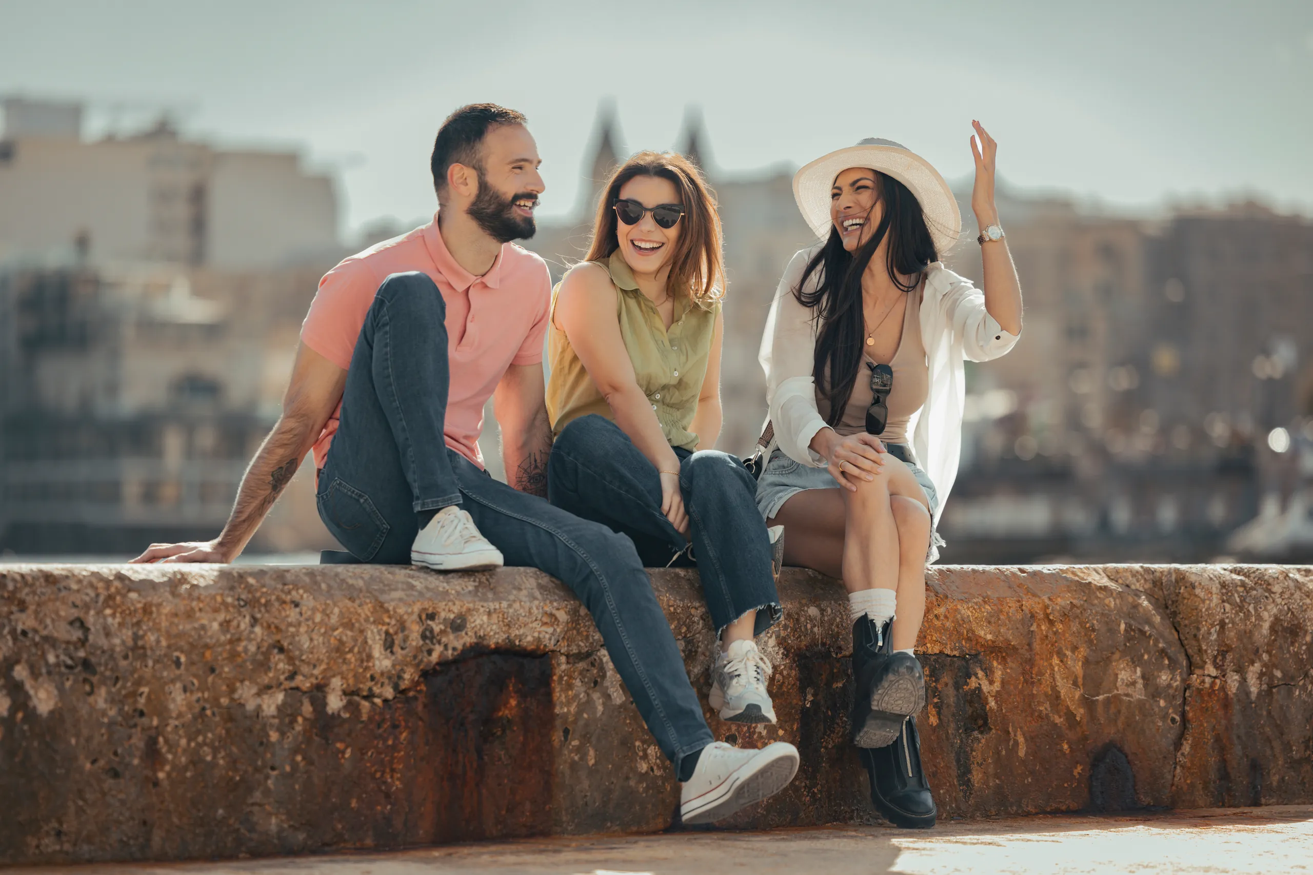 Three friends sitting on a stone sea wall in Malta with the city behind them