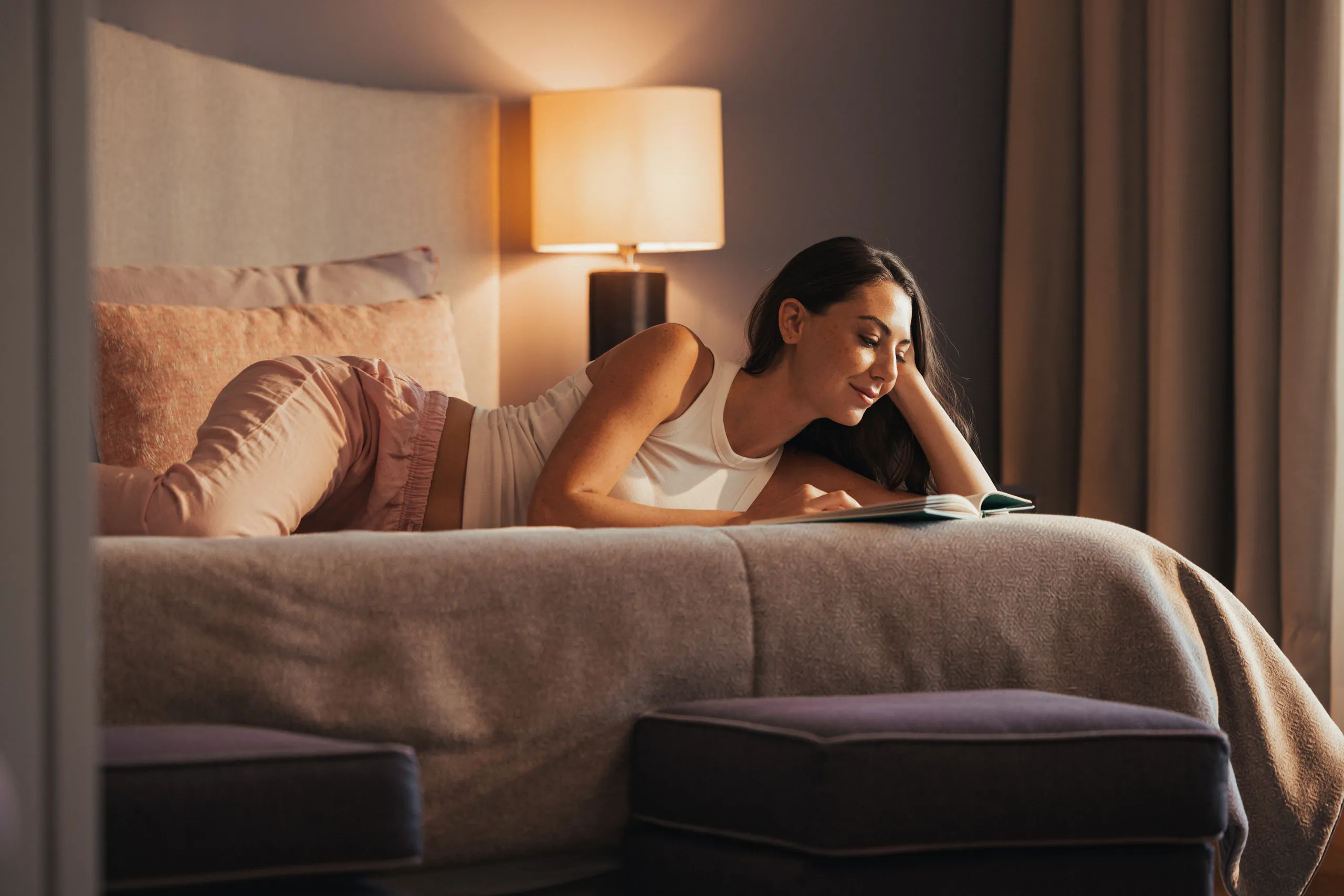Woman lying on a hotel bed reading beside a warm lamp, lifestyle photography for Verdi Hotels
