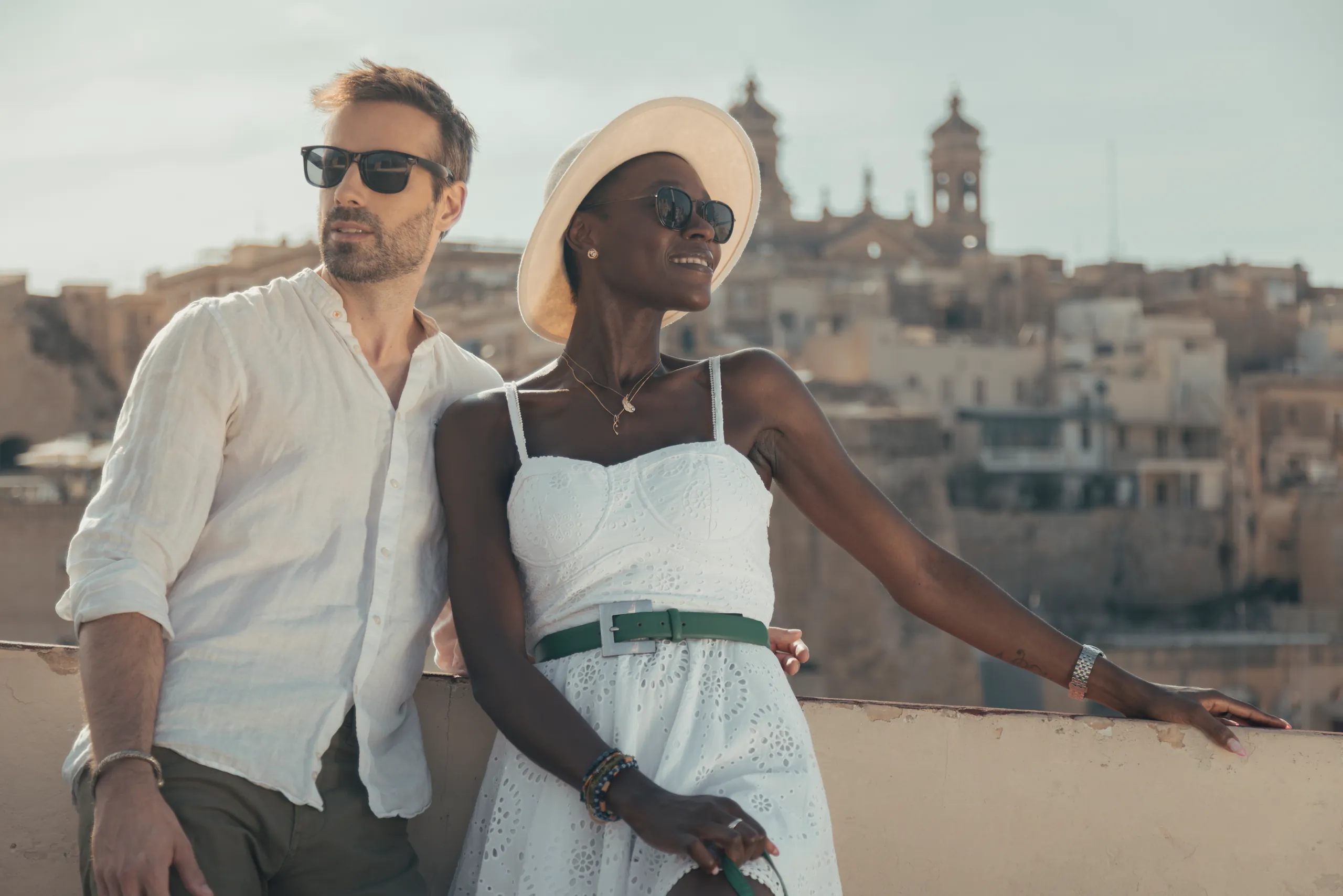 Couple in white outfits leaning on a terrace wall above a hillside city, the woman in a sunhat
