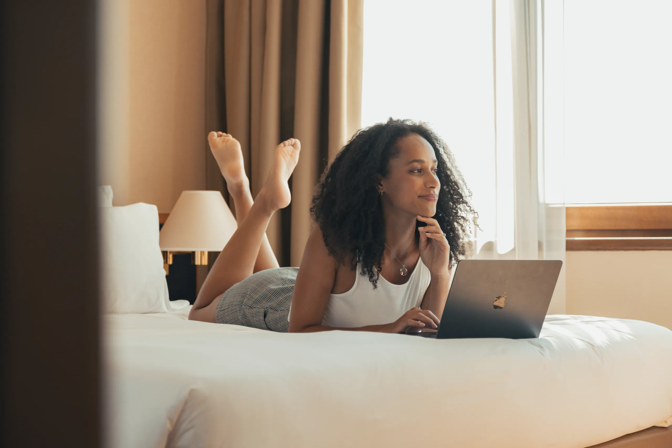 Woman lying on a hotel bed with a laptop beside a window