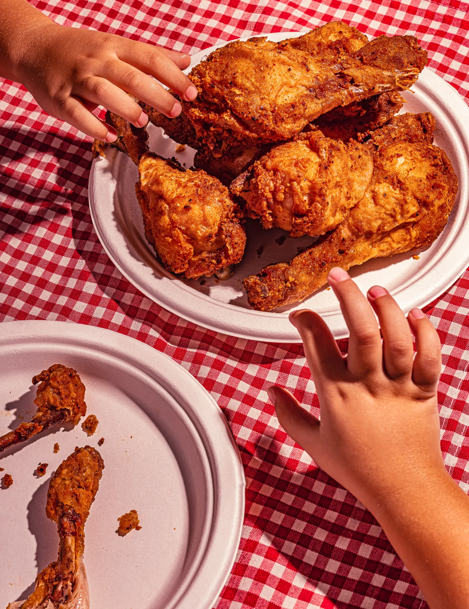Fried chicken on plates on a red gingham cloth, with hands reaching in