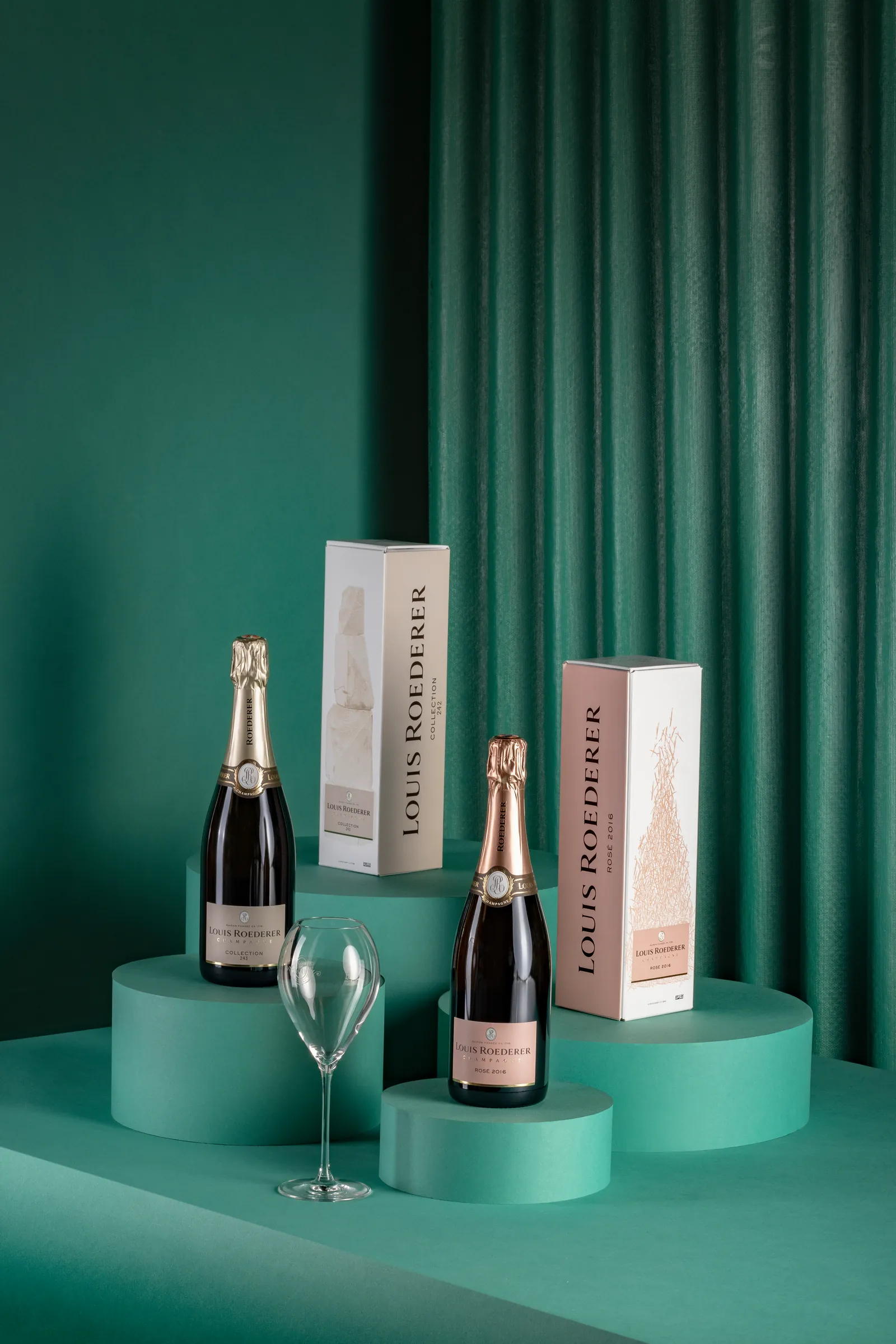 Two champagne bottles with boxes and a glass on stepped teal platforms