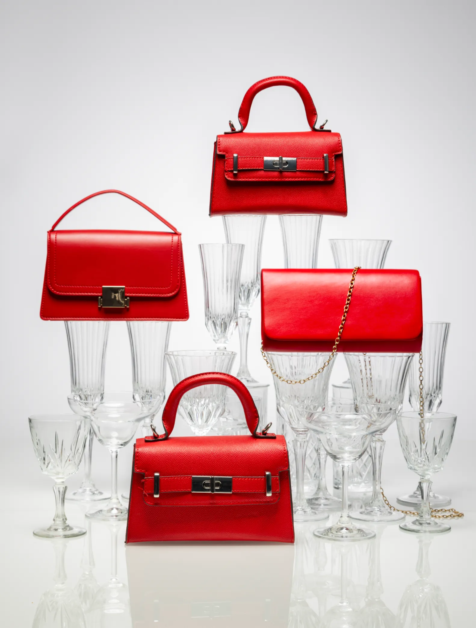 Three red handbags among stemmed crystal glasses on a white background