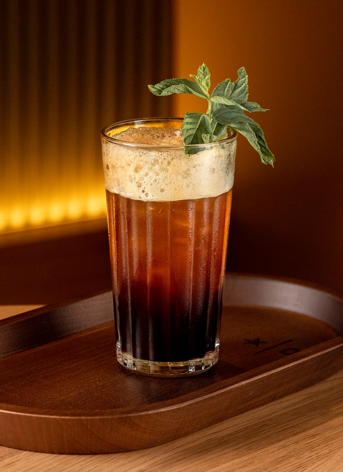 Iced cold brew topped with foam and a sprig of mint in a tall glass on a wooden tray