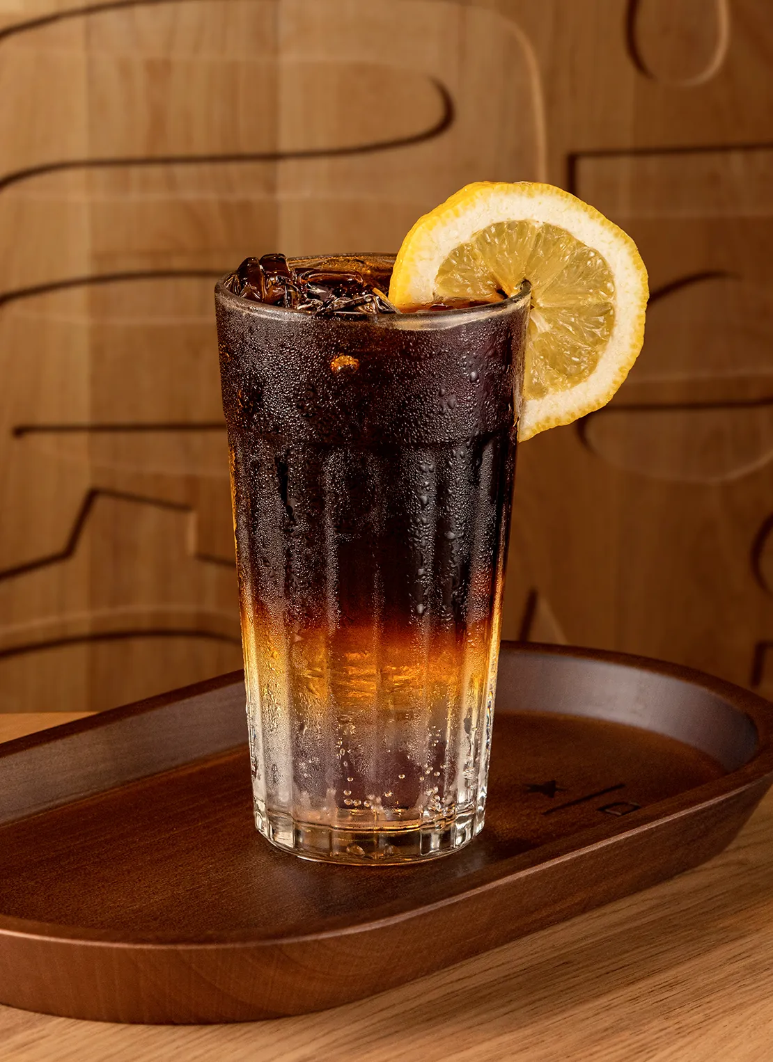 Layered coffee tonic over ice with a lemon slice, served on a wooden tray