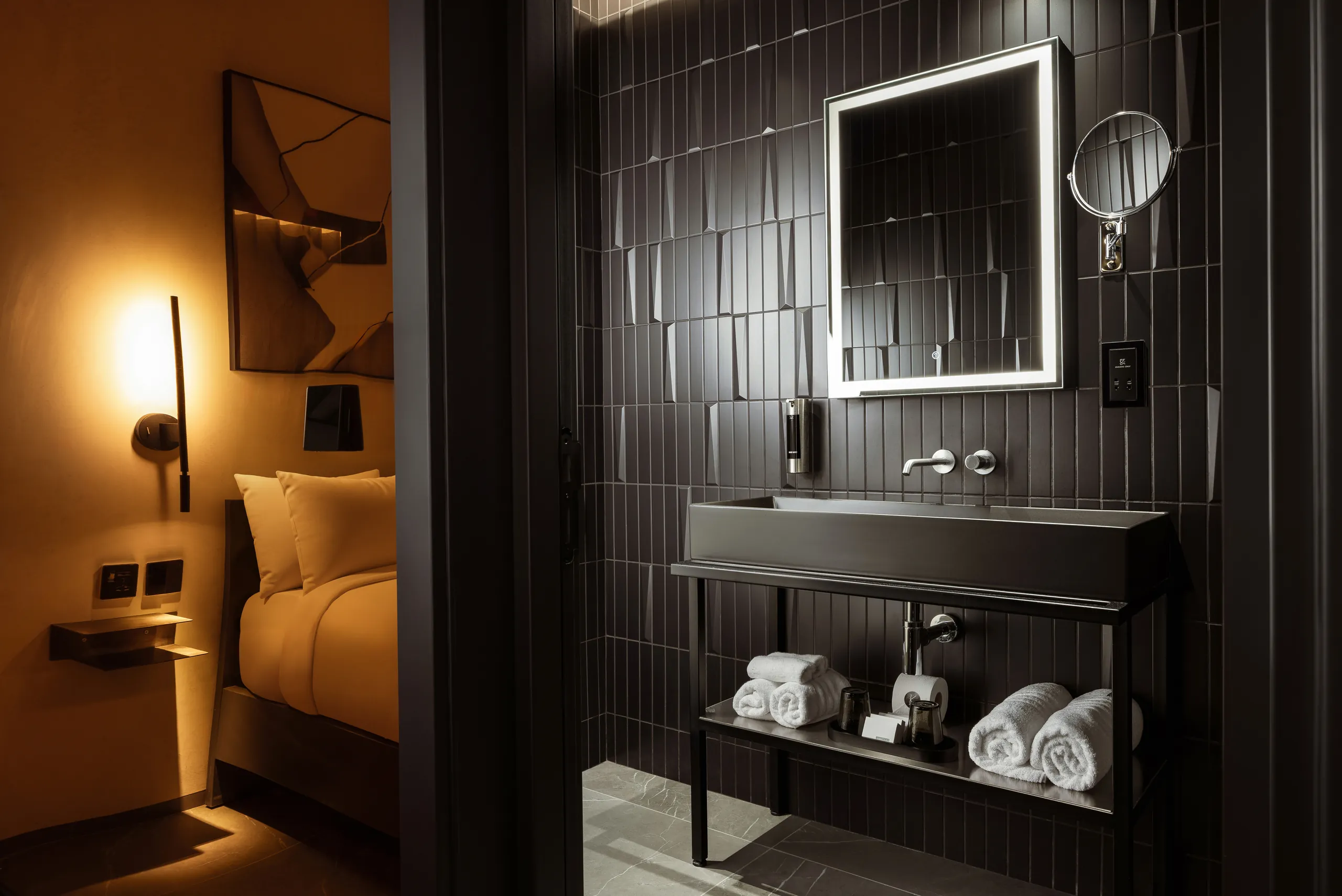 Black-tiled hotel bathroom with a lit mirror, basin and rolled towels