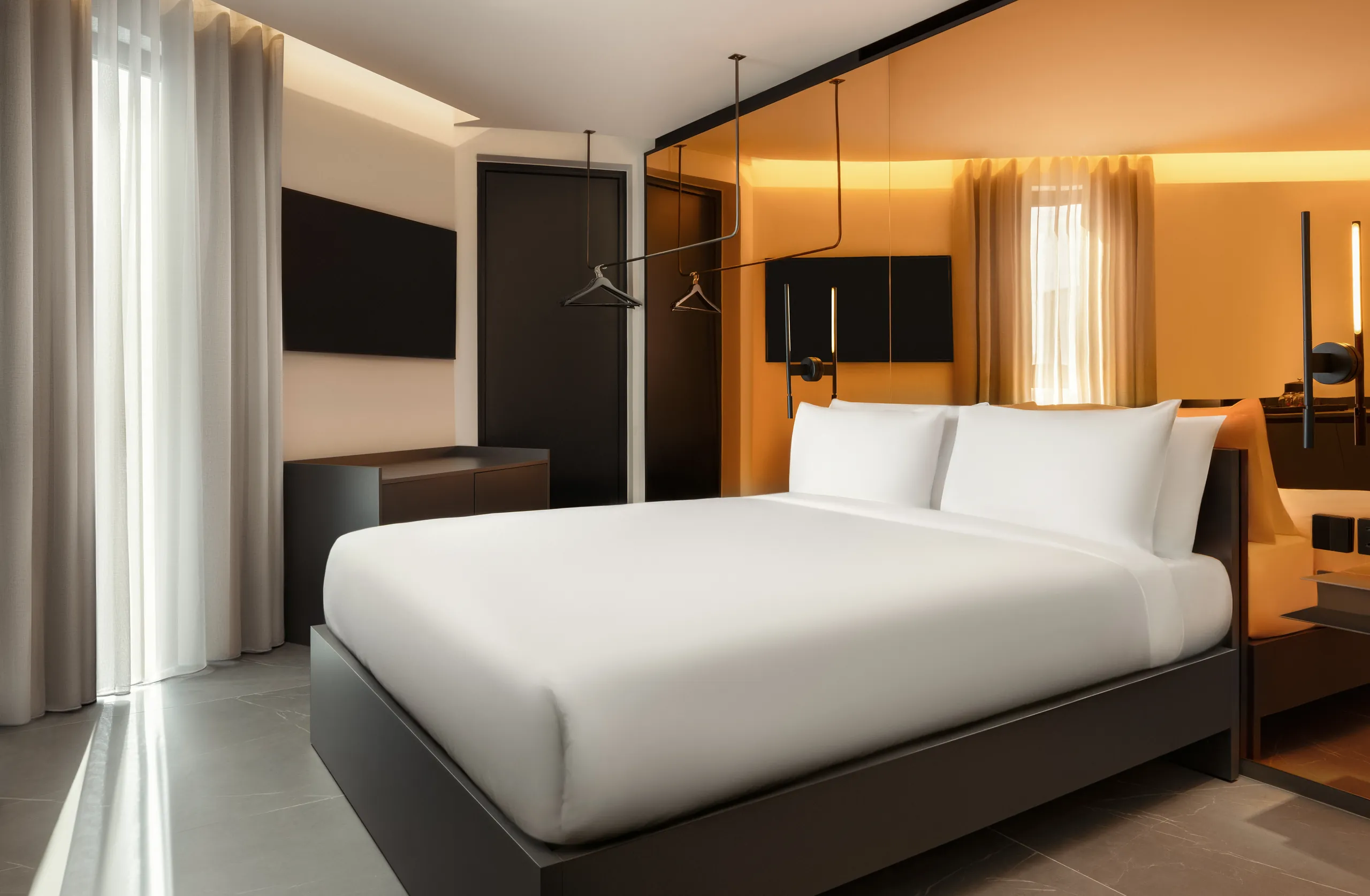 Double hotel room with a white bed, sheer curtains and a wall-mounted TV