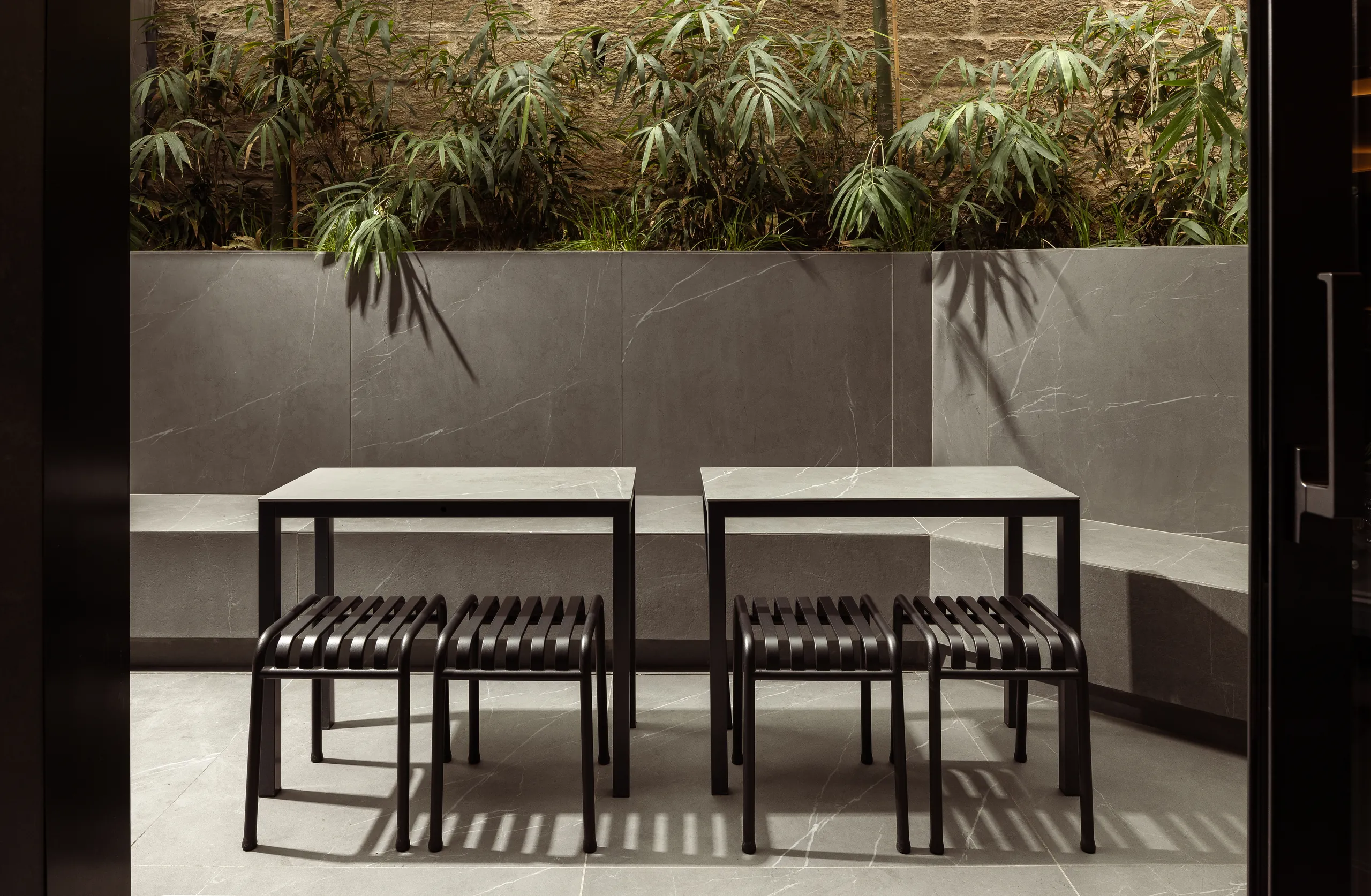 Two outdoor tables with black stools in front of a planted wall